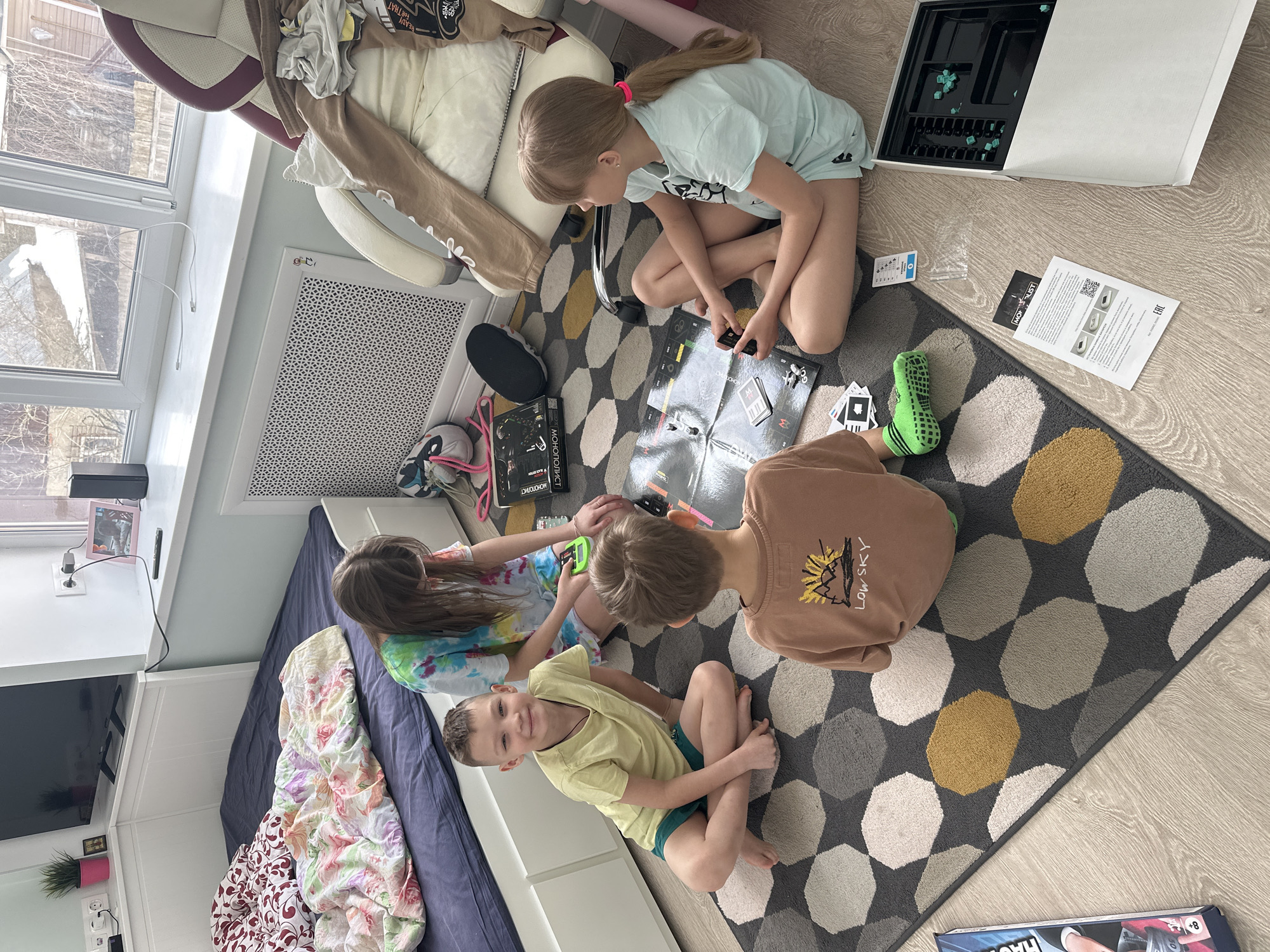 Four children are engaged in a board game on a patterned rug in an indoor setting, likely a home in слобода Старая, Russia. The scene depicts a lively daytime activity, with natural light streaming in from a large window on the left.
The subjects include two girls and two boys, all appearing to be school-aged. They are seated on a dark gray rug with irregular light gray, white, and yellow geometric shapes. They are gathered around a board game, identifiable as "MONOPOLIST" or "MONOPOLY BLACK EDITION," with game cards, green tokens, and the open game box visible on the floor. One girl, with light hair in a ponytail, is intently looking at the game board, holding game pieces. Another girl, with long dark hair, and a boy in a brown "LOW SKY" t-shirt are also focused on the game. A younger boy, wearing a yellow t-shirt, sits cross-legged and smiles towards the camera, conveying a cheerful mood.
The setting appears to be a child's room or a family common area. To the left, a large window reveals a view of bare trees outside, suggesting winter or early spring weather. Below the window is a white radiator. Further left, a bed with a floral patterned duvet and a dark blue sheet is visible, along with a TV on a low white cabinet. Other objects include a white office chair with a beige cover (which has text "READY FOR THAT"), various shoes, wires, and a small potted plant. The flooring is light wood laminate, contrasting with the rug. Instructions for the game, bearing text like "EAC" (a conformity mark) and a QR code, are also scattered on the floor.