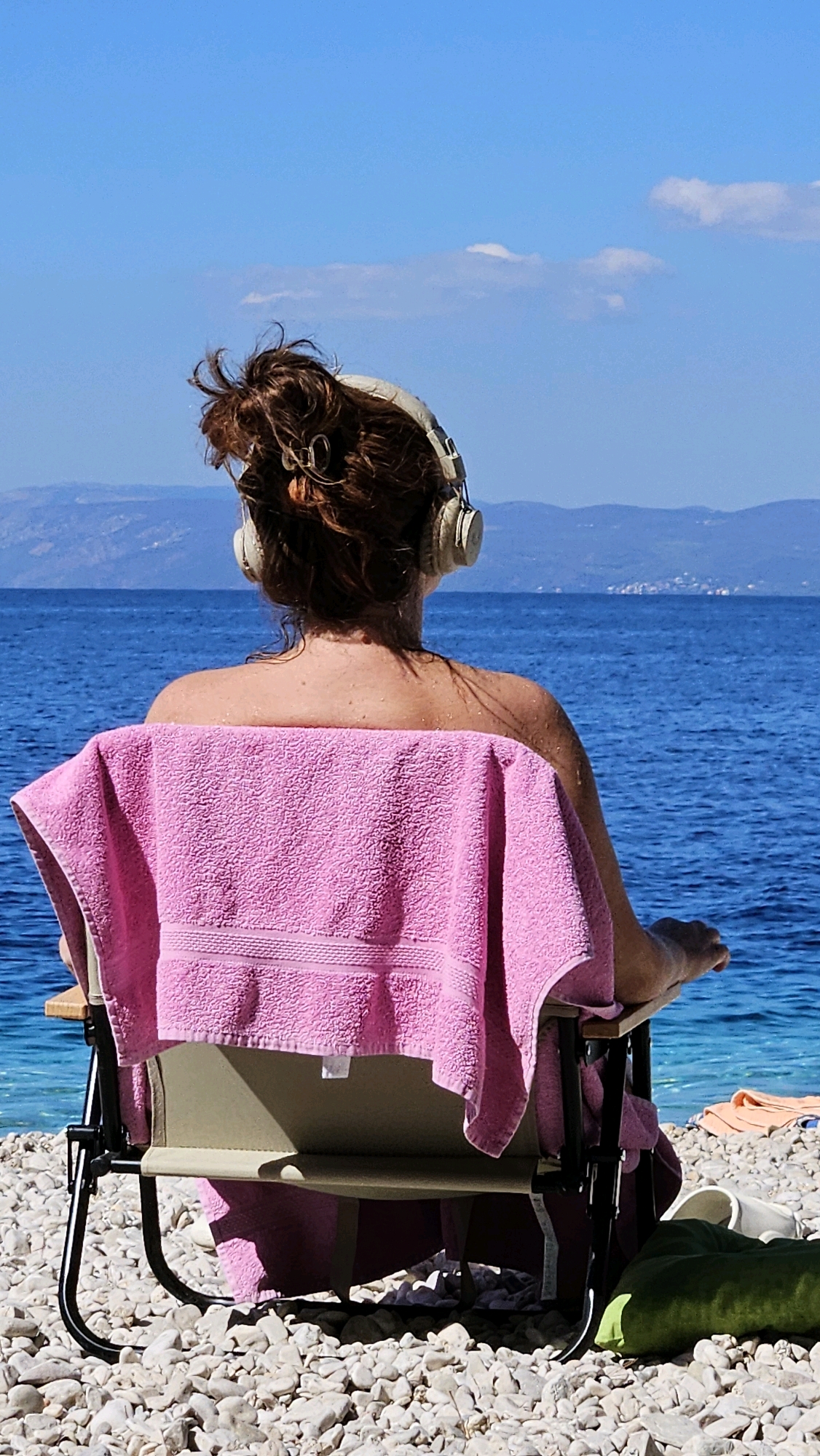 A person wearing headphones sits on a beach chair, facing the ocean. A pink towel is draped over the back of the chair and the person's shoulders. The person's hair is pulled up in a bun. The beach is covered in white pebbles. The water is a deep blue, and mountains are visible in the distance. The sky is clear and blue with a few wispy clouds. The scene appears to be in Općina Podgora, Croatia, likely during daylight hours with bright sunshine. Other beach items, such as another towel and footwear, are visible on the ground near the chair.
