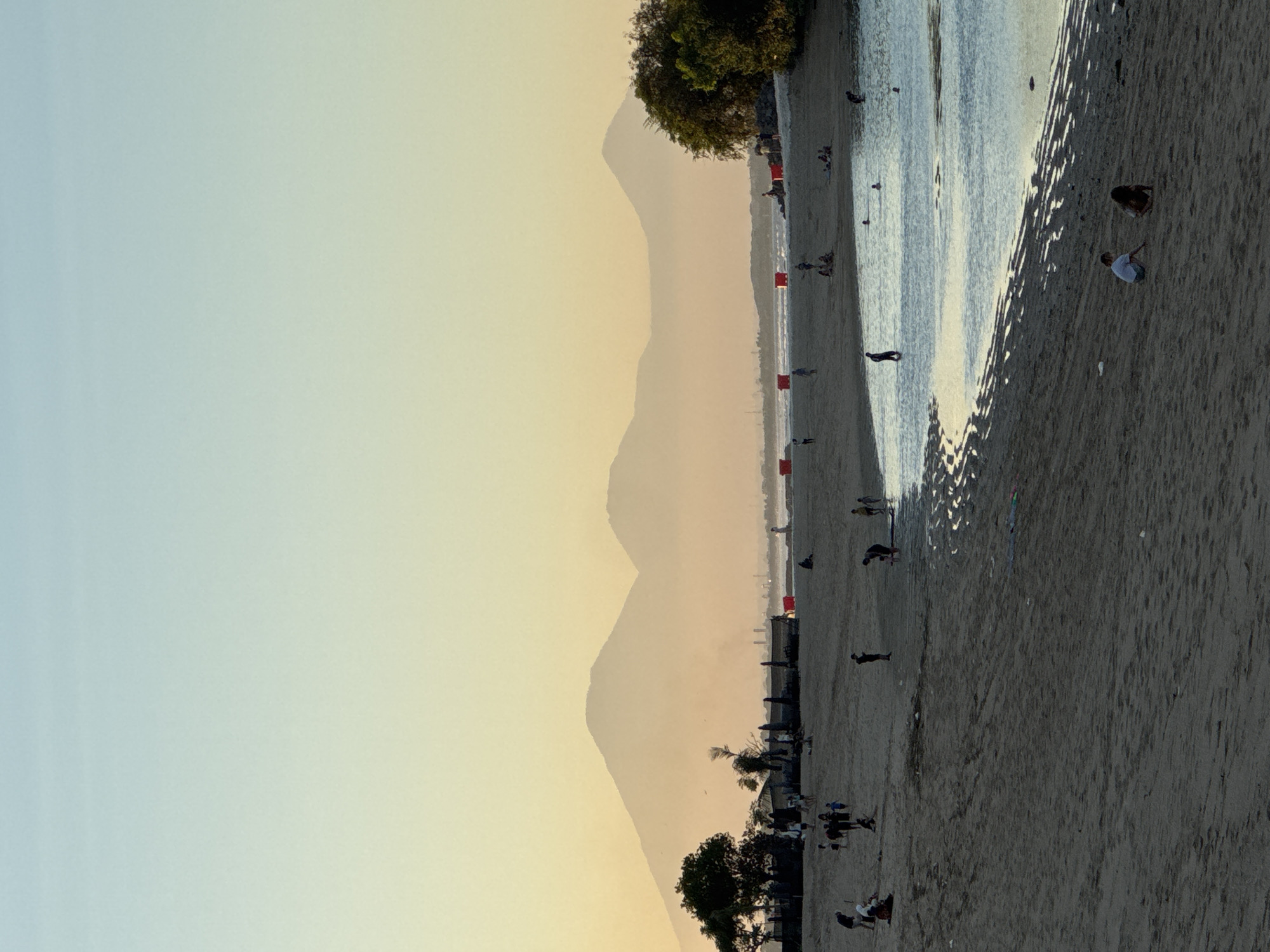 The image shows a beach scene in Kuta Selatan, Indonesia, at sunset. Mountains are visible in the distance, veiled by a warm, pale yellow haze. In the middle ground, the shoreline is marked by red buoys along the beach.
People are scattered across the sandy beach, some near the water and others further inland. Two people are crouched down on the beach. A tree with dark green foliage is at the beginning and end of the shore.
The water appears calm and reflects the sunlight, creating a shimmering effect. The beach itself is mostly light sand, with darker, wet sand closer to the water's edge.