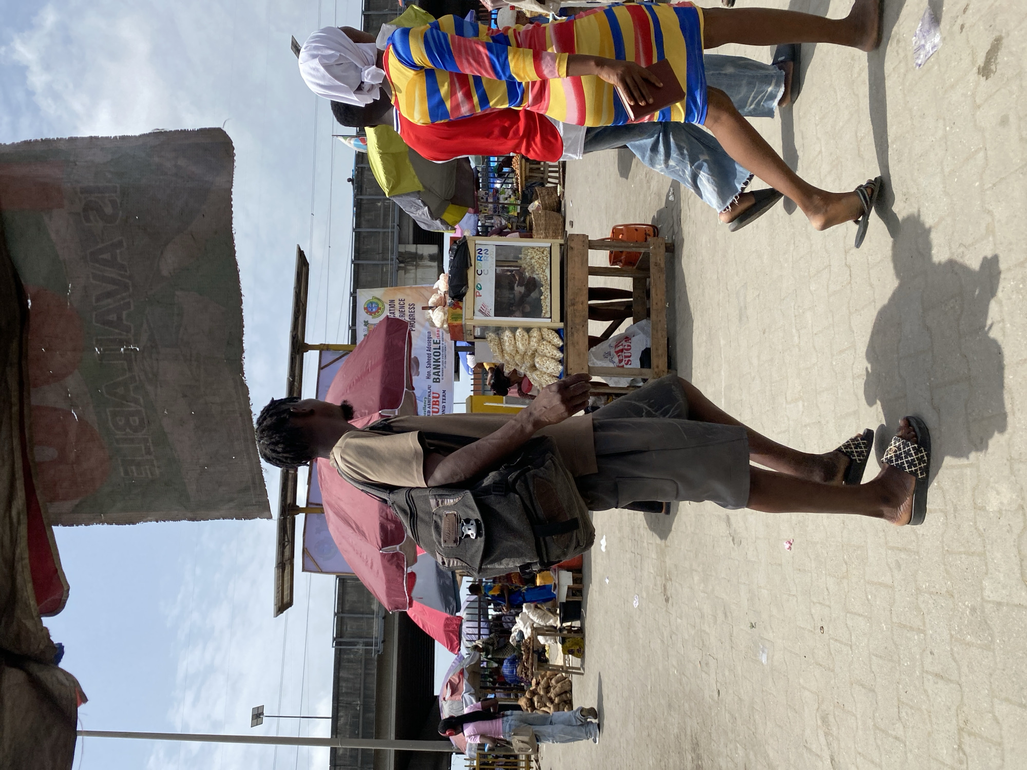 The image shows an outdoor market scene in Lagos, Nigeria. Stalls with goods such as corn and yam line a pathway. People are walking through the market, including a person in a colorful striped dress and a man with a backpack. Signs and awnings are visible above the stalls.
