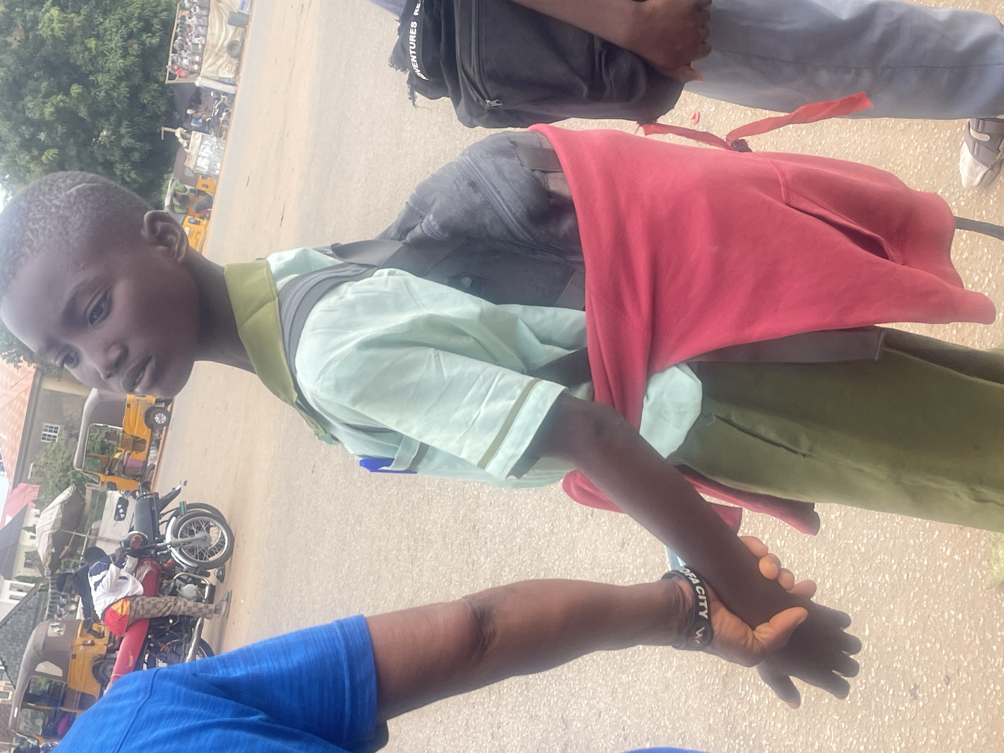 The image shows a young boy wearing a light blue shirt and a dark backpack. He is also wearing a red garment draped over his shoulders, and a green skirt or shorts. He is holding hands with another person whose arm is visible in the foreground, wearing a blue shirt. The boy appears to be looking to his left with a neutral expression. The scene is outdoors, with a dusty road and several vehicles in the background, including a motorcycle and two yellow auto-rickshaws. There is also some greenery and buildings visible. The overall lighting suggests it is daytime. A black backpack with the word "ADVENTURES" is visible at the top of the image. Another person's legs are also visible, wearing grey trousers and white sneakers. A black wristband on the hand holding the boy's hand has "OFA CITY W" written on it. The context provided indicates the location as Galadimawa Village, Nigeria.