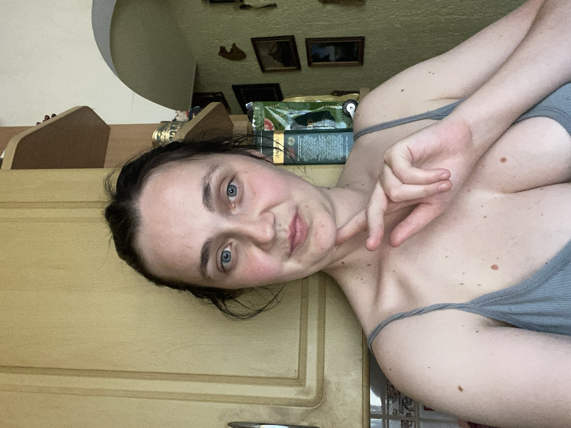A young woman with dark hair and blue eyes is lying down, with her head tilted to the side and her right hand near her face. She is wearing a grey tank top. Behind her, there are several framed pictures on the wall and a stack of what appear to be packages of tea or coffee. The background also includes kitchen cabinets. The lighting suggests it is daytime, though the exact time is not discernible. The woman's expression appears to be neutral or slightly contemplative.
