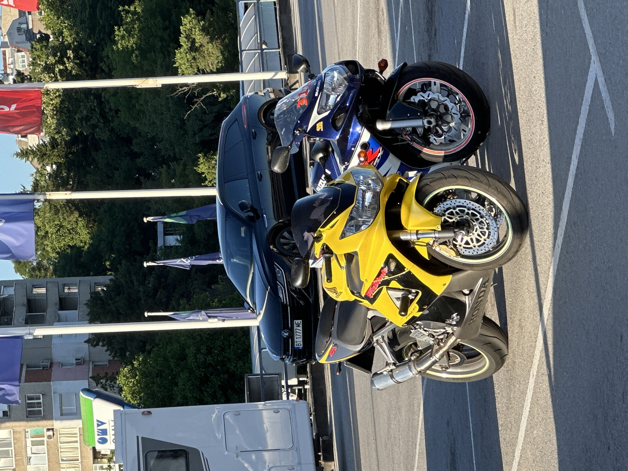 In Veliko Tarnovo, Bulgaria, two motorcycles are parked in a lot. The motorcycle in the foreground is yellow and black, with the "HONDA" logo visible on its side. Behind it is a blue and white motorcycle, with the "SUZUKI" logo on its fairing. Both motorcycles are sports bikes. In the background, a black Volkswagen Arteon station wagon is parked, displaying a license plate that reads "BT 1777 ME". A white recreational vehicle is also parked to the left of the frame. The scene is set against a backdrop of trees and buildings, with several flags on poles visible. The ground is paved asphalt with parking lines. The lighting suggests it is daytime, with strong shadows cast on the ground.