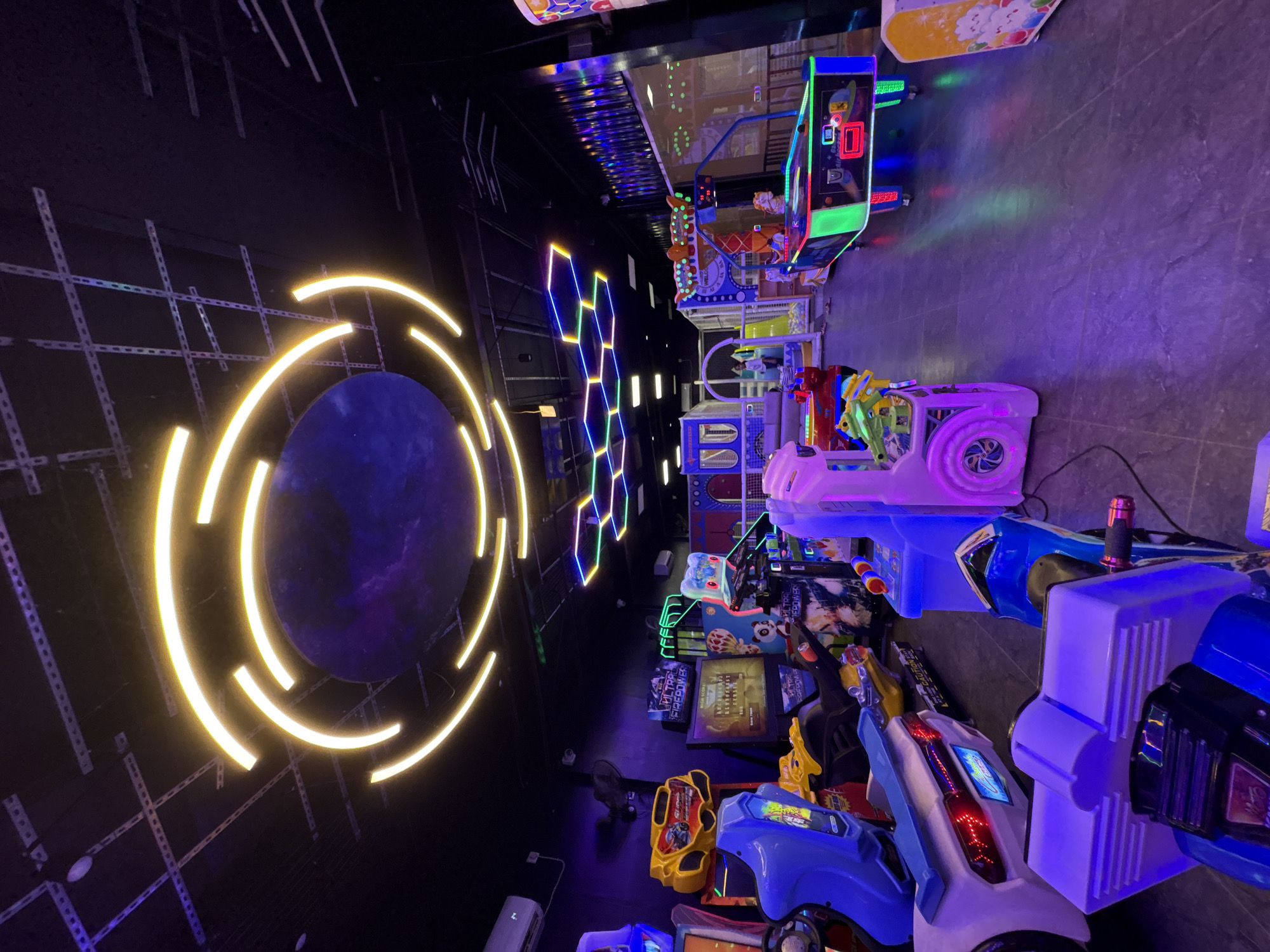 The image depicts an indoor arcade or entertainment center, characterized by its dark ambiance illuminated by vibrant neon and LED lights. The scene is dominated by various arcade games arranged in rows. In the foreground and extending to the right, several shooting games are visible, featuring colorful housings in blue, white, and purple, equipped with light-up gun controllers. Further back, other game machines, possibly including racing games, can be seen with their glowing screens and interactive setups.
The left side of the frame showcases prominent decorative lighting. A large, dark, circular object, possibly representing a planet or portal, is encircled by bright, glowing yellow curved light strips that create a dynamic visual effect. Above and to the right of this, a pattern of interconnected hexagonal light fixtures glows in various colors like blue, green, and purple, contributing to a futuristic aesthetic. The floor is a dark, hard surface, reflecting some of the colorful lights from the games and wall installations. The ceiling is dimly lit, revealing some structural elements.
There are no people visible in the image, implying the area might be empty or photographed during off-peak hours. The arrangement of numerous game machines clearly indicates a setting for gaming and entertainment activities. The overall atmosphere is created by the low light conditions and the strong contrast of the colorful, glowing elements.
Visible text includes "ULTRA FIREPOWER" prominently displayed on one of the game screens, and "HIGH SCORE" on another screen, along with other unreadable game-related text. Although the context provided is Chennai, India, there are no specific visual cues within the image that uniquely identify this location.