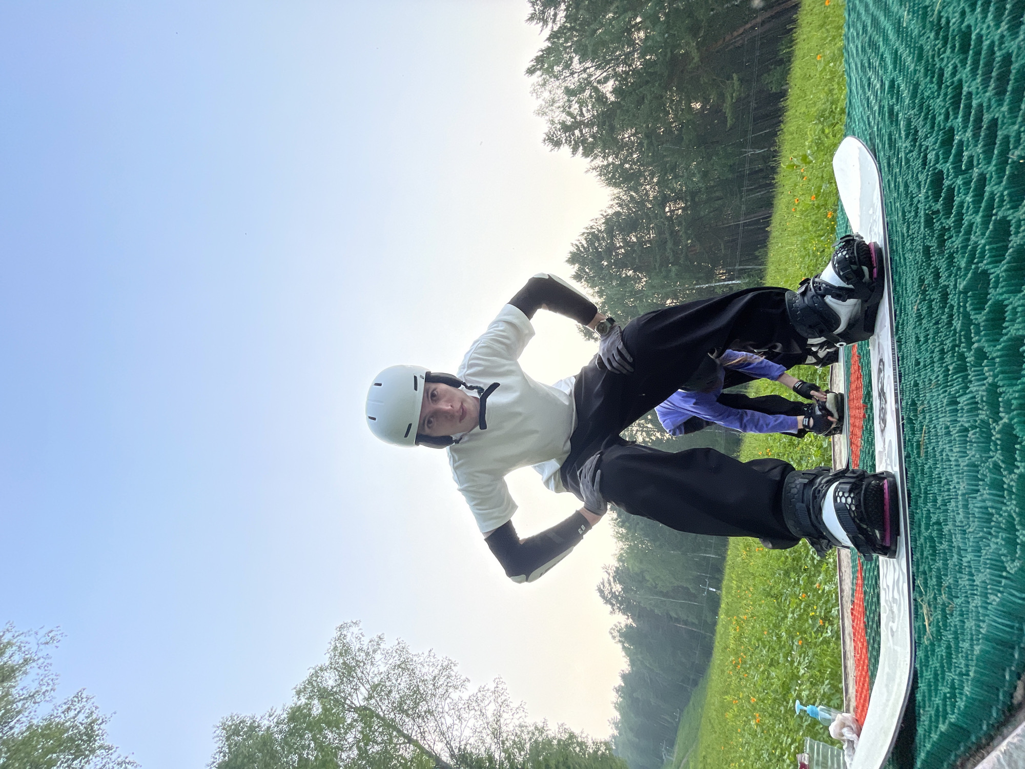 A low-angle, full shot captures a young person in a snowboarding stance on a green mat, with a snowboard beneath their feet. The person is wearing a white helmet, a white t-shirt, black pants, and grey gloves, with elbow pads visible on their arms. Their stance is wide, with knees bent and hands on their hips. In the background, a grassy slope dotted with yellow flowers leads up to a dense forest. The sky is clear and pale blue, with the soft light suggesting early morning or late evening. The location is the Divnogorsk Ski Resort in Divnogorsk, Russia, a place known for its natural beauty and outdoor recreational activities.