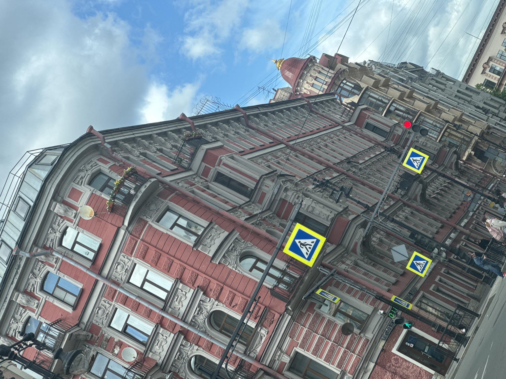 The image depicts a street scene in Saint Petersburg, Russia, showcasing the architectural details of a prominent building. The building is characterized by its ornate façade, featuring red and grey brickwork, intricate molding, and multiple windows. The architecture suggests a historical or classical design. Visible above the building are elements of the sky, including blue patches and scattered clouds. Street elements visible include traffic lights, pedestrian crossing signs, and utility wires overhead. Sidewalk with some people is also noticeable. The perspective of the shot is angled upwards, emphasizing the height and grandeur of the building.