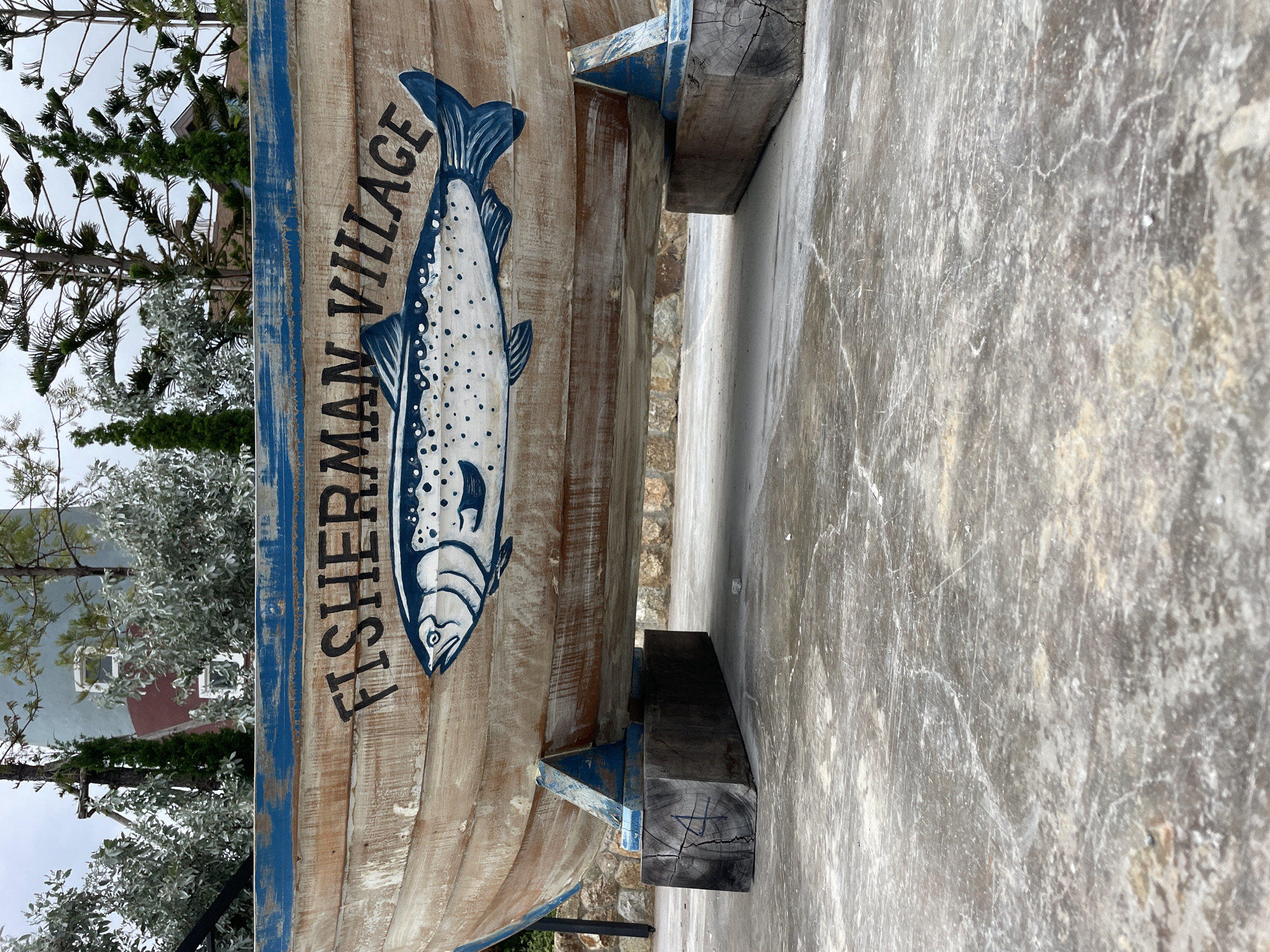 A vintage wooden boat, painted with blue trim and a stylized fish, rests on wooden blocks in what appears to be an outdoor seating area in Pattaya City, Thailand. The boat is adorned with the words "FISHERMAN VILLAGE" in black lettering. Behind the boat, a building with blue and red sections is visible, surrounded by various trees and foliage. The ground is paved with concrete, showing signs of wear and cracks. The sky is overcast.