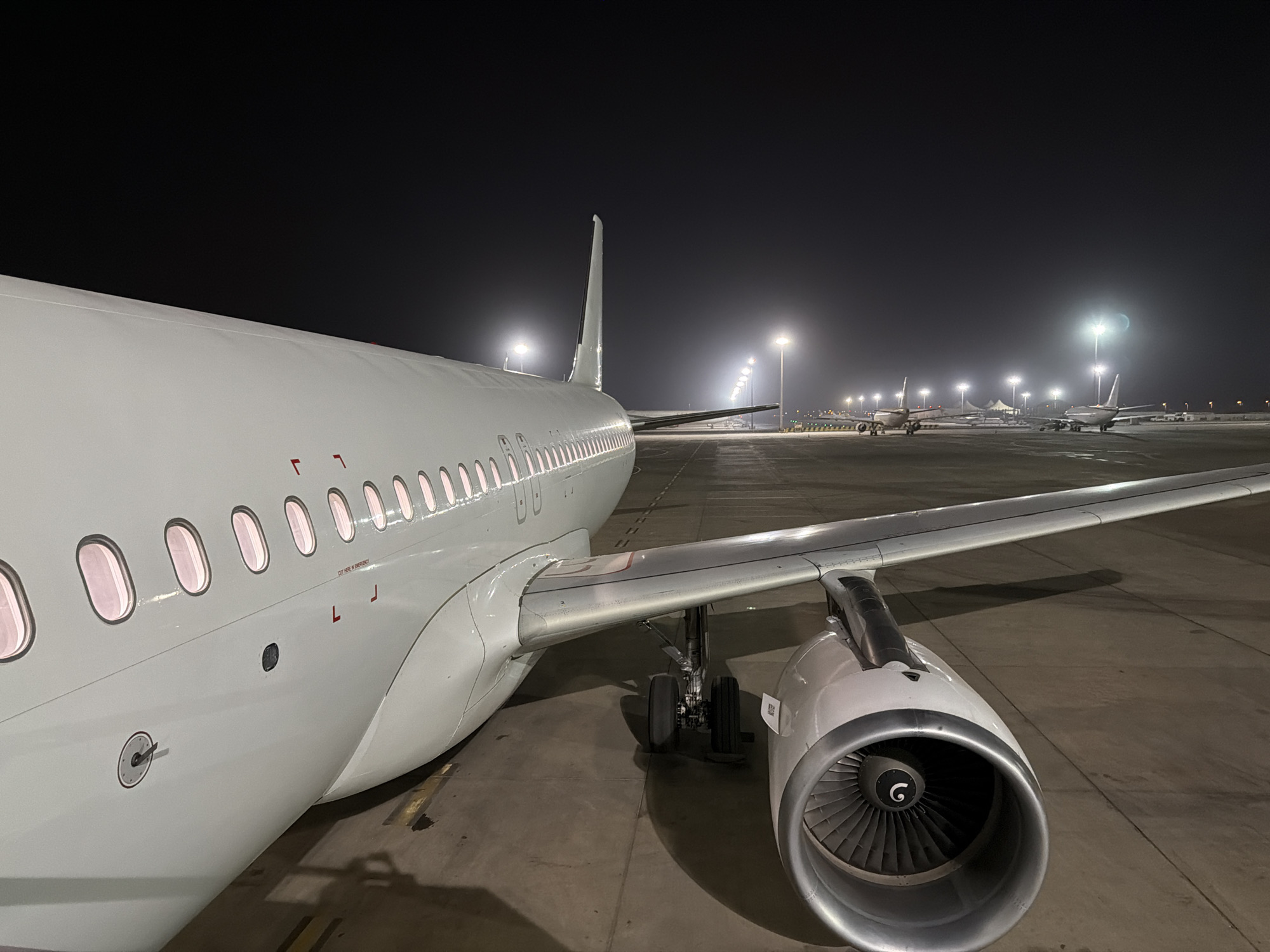 At night in what appears to be Blaise Diagne International Airport in the city of Communauté rurale de Diass, Senegal, an airplane, likely an Airbus A320 family aircraft, is parked on the tarmac. It is white, with visible windows, a wing, and one engine. Other aircraft are visible further in the distance under bright runway lights. The ground is concrete. The sky is dark.