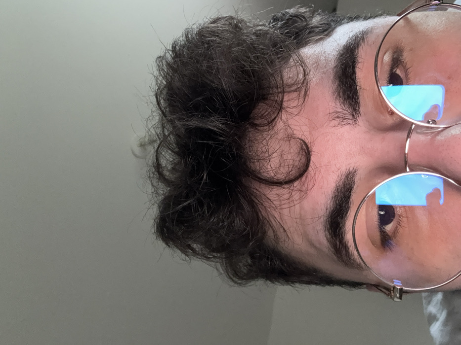 The scene is located in Rennes, France. The image provides a close-up, low-angle view of a person's upper face. The subject has dark, curly hair styled above the forehead and prominent dark eyebrows. The individual is wearing round, gold-rimmed eyeglasses. The lenses of the eyeglasses exhibit distinct blue, rectangular reflections, consistent with light emitted from a digital screen. The subject's eyes are partially visible behind the lenses. The background features a plain, light-colored ceiling. A circular, white ceiling fixture or detector is partially visible above and behind the subject's hair, in the upper right quadrant of the image. A minimal portion of light-colored fabric, likely clothing, is visible at the extreme bottom left.