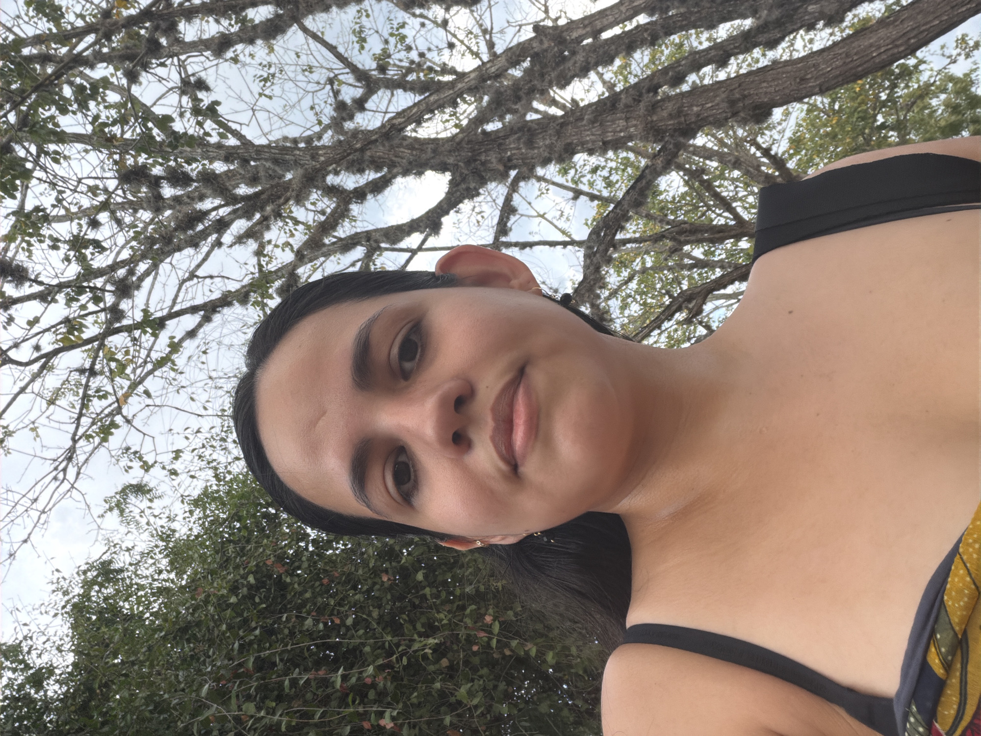 A woman with dark hair and light skin is in the foreground of the image, looking slightly up at the camera. She is wearing a black bra or swimsuit top and part of a colorful garment can be seen on the right. The background shows trees with bare branches and some green foliage, suggesting a natural outdoor setting. The sky is overcast, with a pale, cloudy appearance. The image appears to have been taken from a low angle, looking up at the subject and the trees. The location in the city of Ciudad Guayana, Venezuela, is not evident from the image.