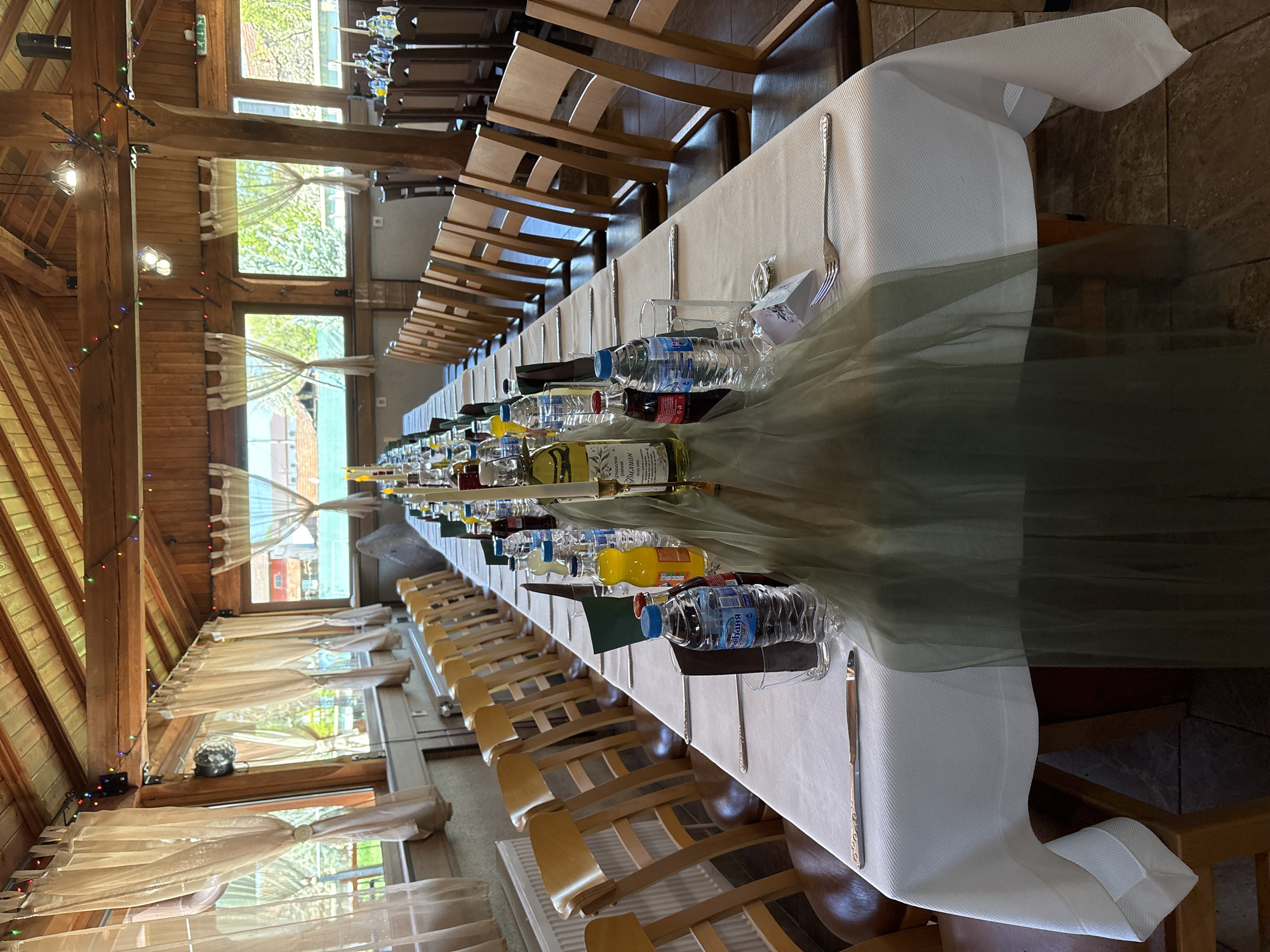 The image shows a long, set dining table at an event likely taking place indoors in Kopilovtsi, Bulgaria. The table, covered with a white linen tablecloth and a semi-transparent green runner, is adorned with glassware, utensils (forks and knives), bottled drinks (water, juice, and possibly wine), and small gift boxes placed at each setting. The rows of chairs indicate a formal seating arrangement. The location appears to be a restaurant or event space with wooden beams and supports that are indicative of an indoor location. The location is defined by the presence of curtains hung on the windows facing outside.