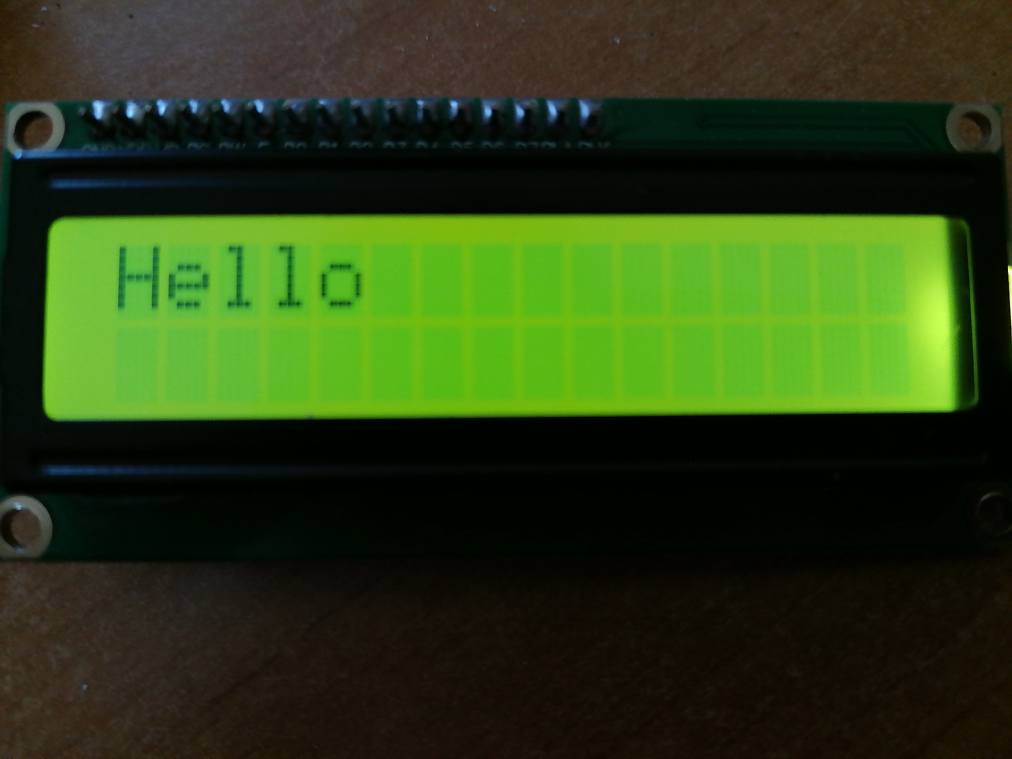 A close-up shot shows a green LCD screen displaying the word "Hello". The screen is part of a circuit board with a row of pins along the top. The circuit board is mounted on a black plastic casing. The background is a brown, wooden surface.