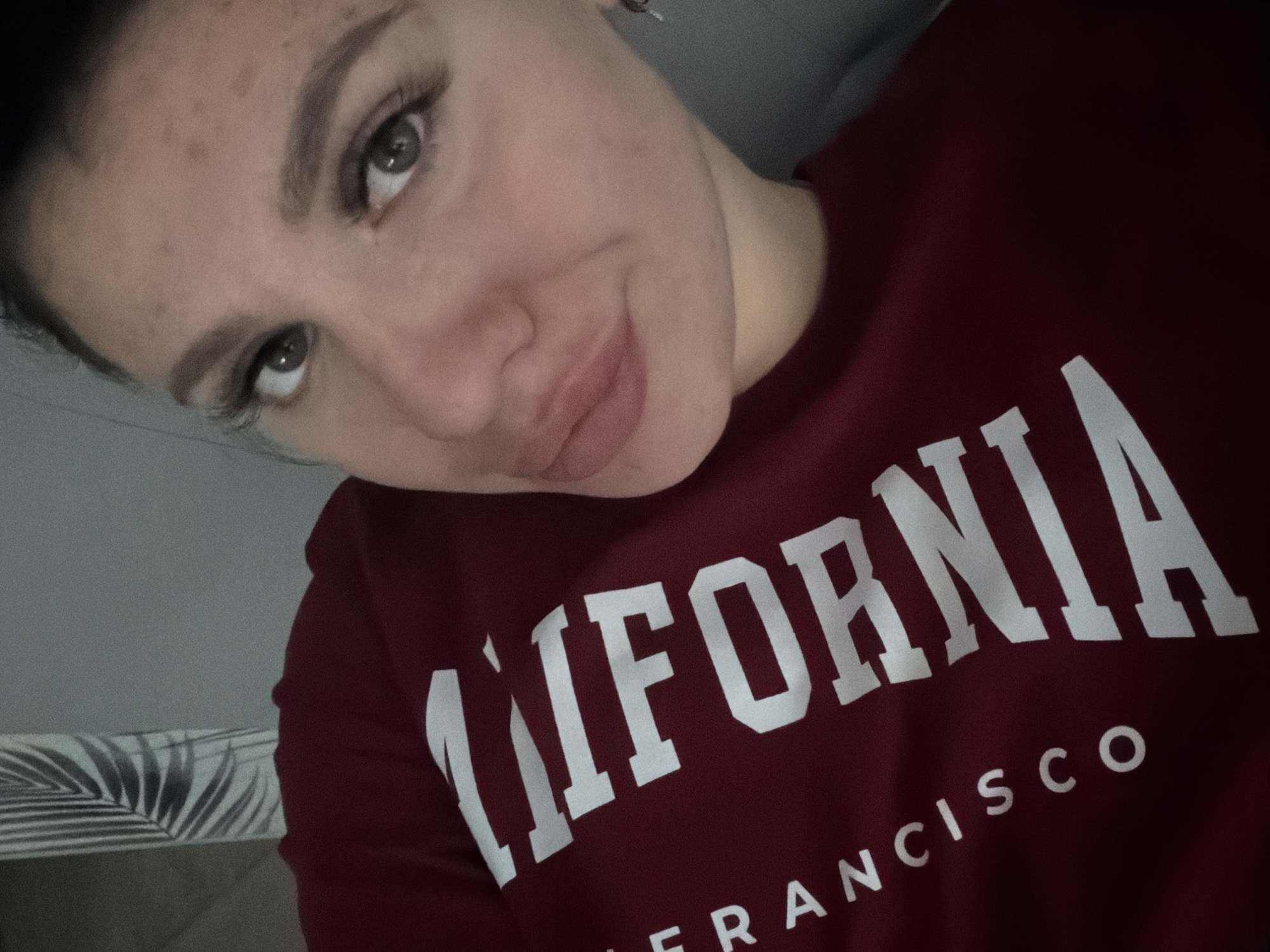 A close-up, angled selfie of a young woman with brown hair pulled back and dark eyebrows. She is wearing a maroon sweatshirt with the words "CALIFORNIA" and "FRANCISCO" printed in white block letters. Her facial features include light-colored eyes, full lips, and visible eyelashes. The background is blurred, showing a gray wall and a hint of a decorative leaf pattern on a vertical surface. No specific location in Fokino, Russia, is identifiable from the image.