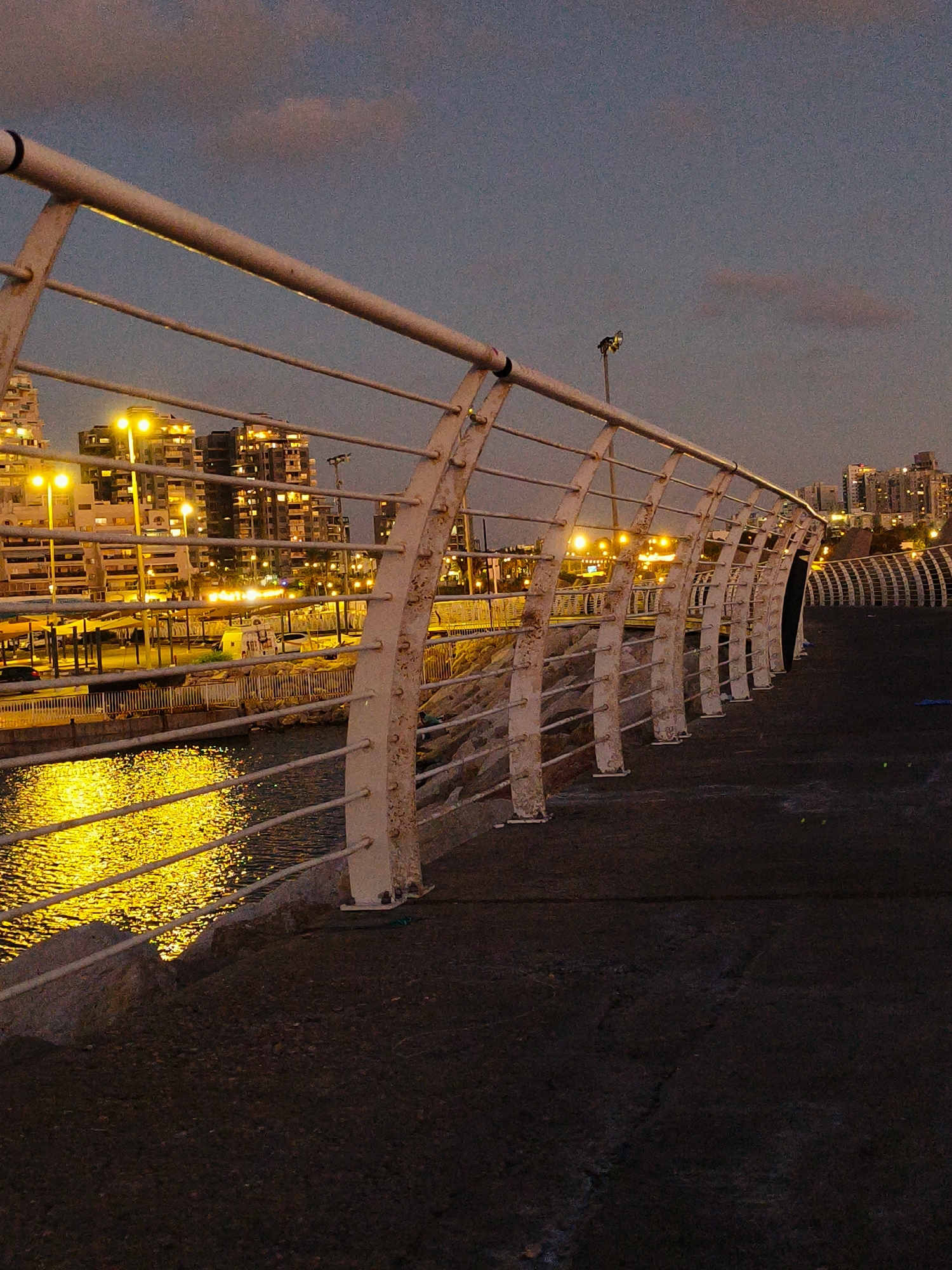 The image depicts a waterfront promenade in Ashkelon, Israel, during twilight. A white metal railing, showing signs of wear and rust, lines the edge of the promenade, creating a series of arches. Beyond the railing, calm water reflects the warm glow of lights from buildings in the background, suggesting a city setting. These buildings, appearing as a cluster of high-rise structures, are illuminated with warm yellow lights, indicating that it is evening. The sky above is a dusky blue with subtle hints of pink, characteristic of sunset or pre-dawn hours. The promenade itself is paved with dark asphalt, showing some texture and wear. The overall atmosphere is serene, with the urban landscape and the reflective water creating a peaceful, yet lively, scene.