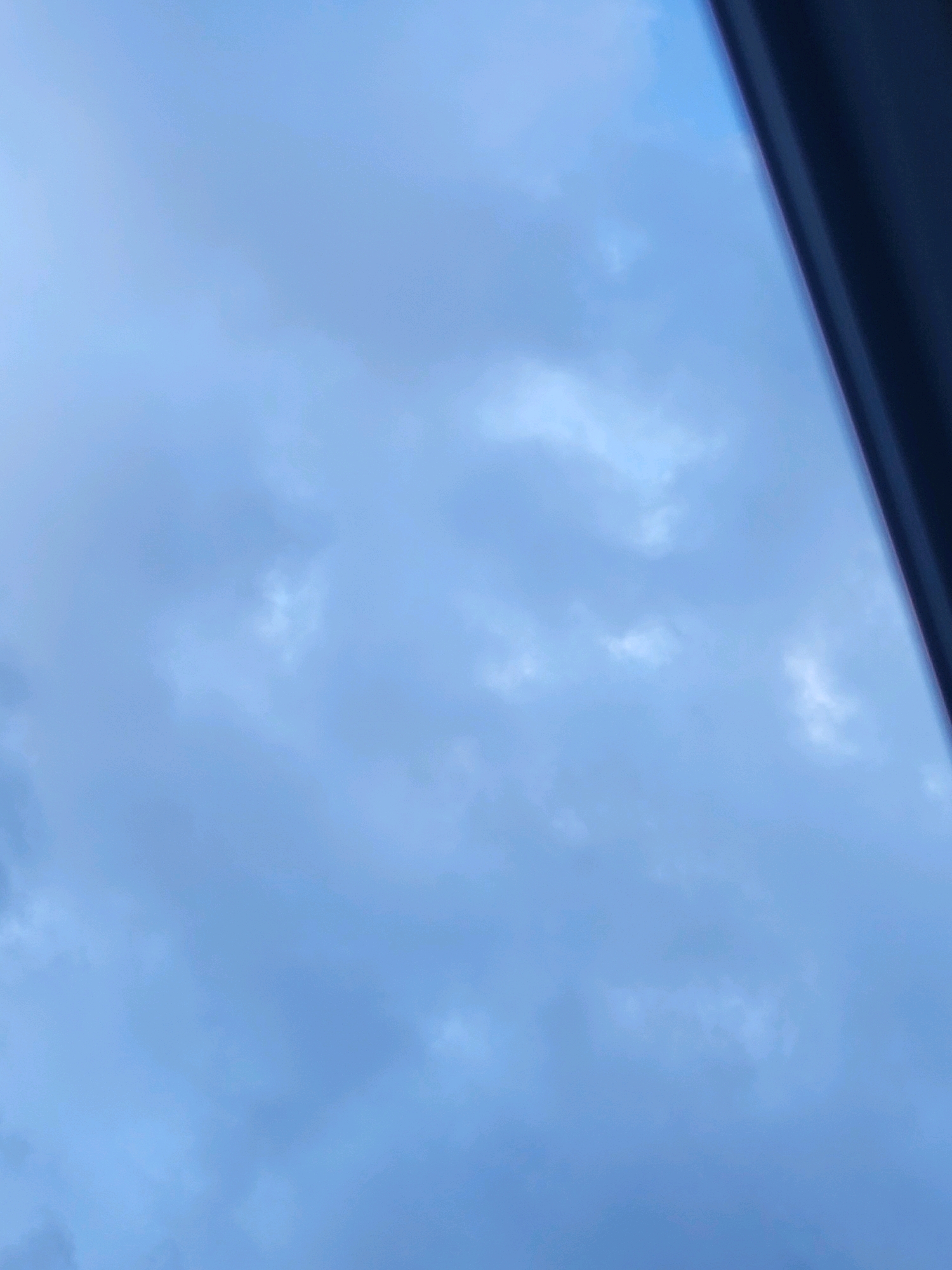 The image shows a cloudy sky with a dark object on the right side, possibly a window frame or a building edge, obscuring a portion of the view. The sky is predominantly blue with varying shades of gray and white clouds scattered throughout. The lighting suggests it is daytime, likely overcast. There are no people or animals visible in the image. The scene is entirely composed of the sky and the obstructing dark object. There is no information to determine the location in Jelgava, Latvia.