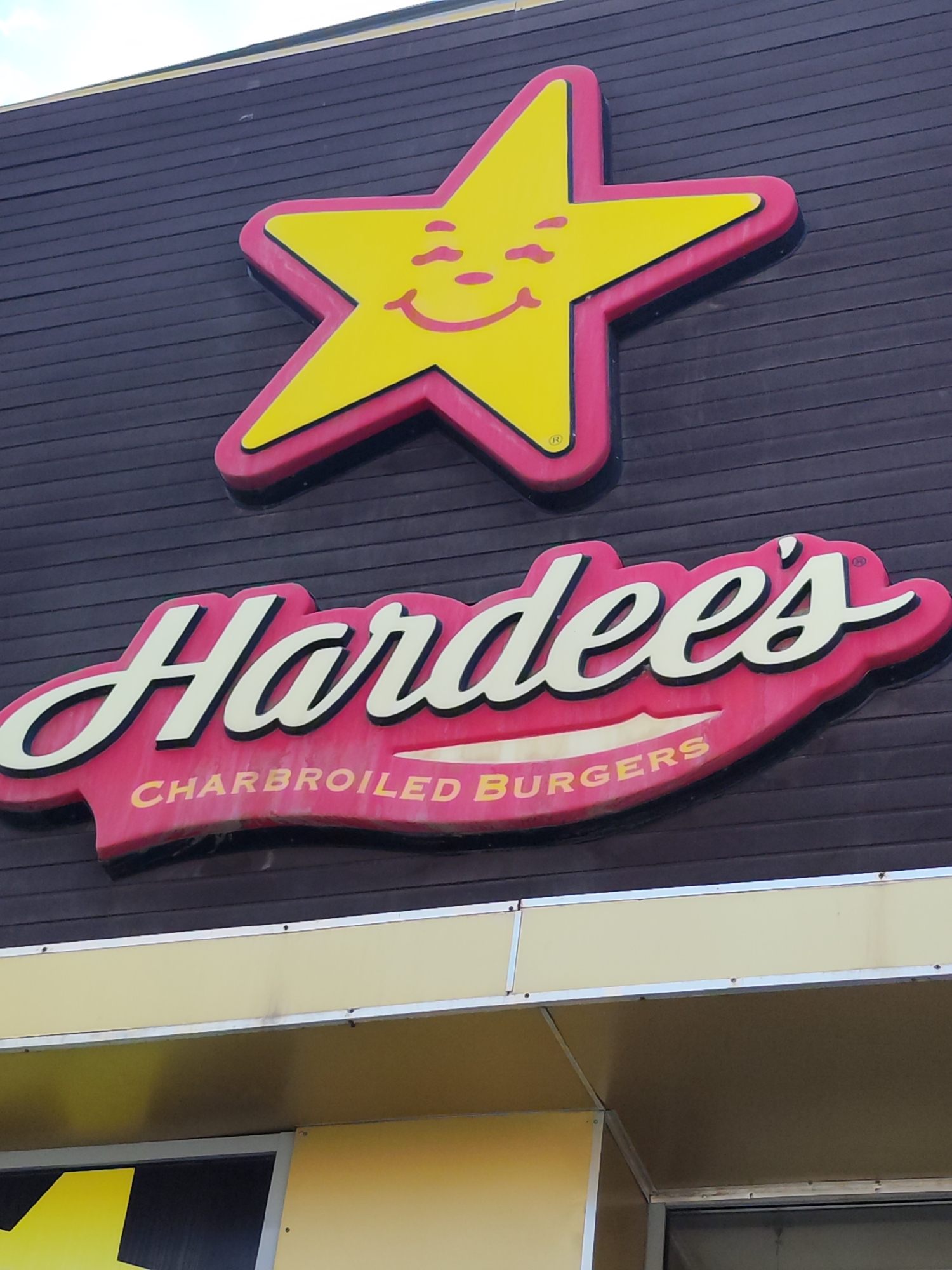 This image shows the exterior of a Hardee's restaurant, a fast-food chain. The dominant elements are the Hardee's logo, which features a yellow star with a smiley face, and the brand name "Hardee's" written in white script on a red background, accompanied by the tagline "CHARBROILED BURGERS." The building facade is dark brown with horizontal paneling. Below the main signage, there's a yellow structure with metal trim, and partially visible in the lower part of the image are entrances and further yellow panels, suggesting the restaurant is open or accessible. There are no people or vehicles visible in the frame. The image appears to have been taken outdoors, under daylight conditions, with a slight upward angle. The specific location in Astana, Kazakhstan is not indicated by any visible markers in the image itself.