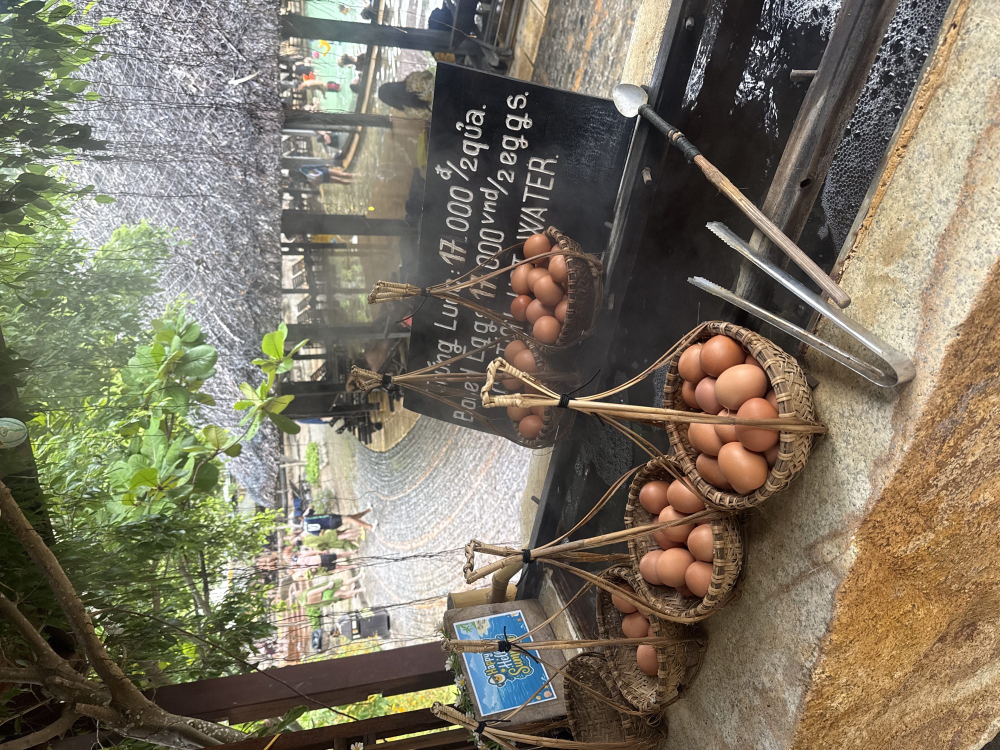 This image depicts an outdoor food stall at a hot spring resort, likely in Nha Trang, Vietnam, offering boiled eggs cooked in hot water. In the foreground, several woven baskets filled with brown eggs are displayed on a stone counter. Behind the baskets, a dark chalkboard displays pricing and product information in white lettering, indicating "Boiled Eggs" and "HOT WATER." Tongs and a ladle rest near a trough filled with bubbling hot water, suggesting the eggs are prepared on-site. In the background, blurred figures of people are visible enjoying the hot spring facilities, with thatched-roof structures and lush greenery forming the resort's ambiance. The overall scene captures a rustic and natural setting where visitors can experience a local culinary tradition.
