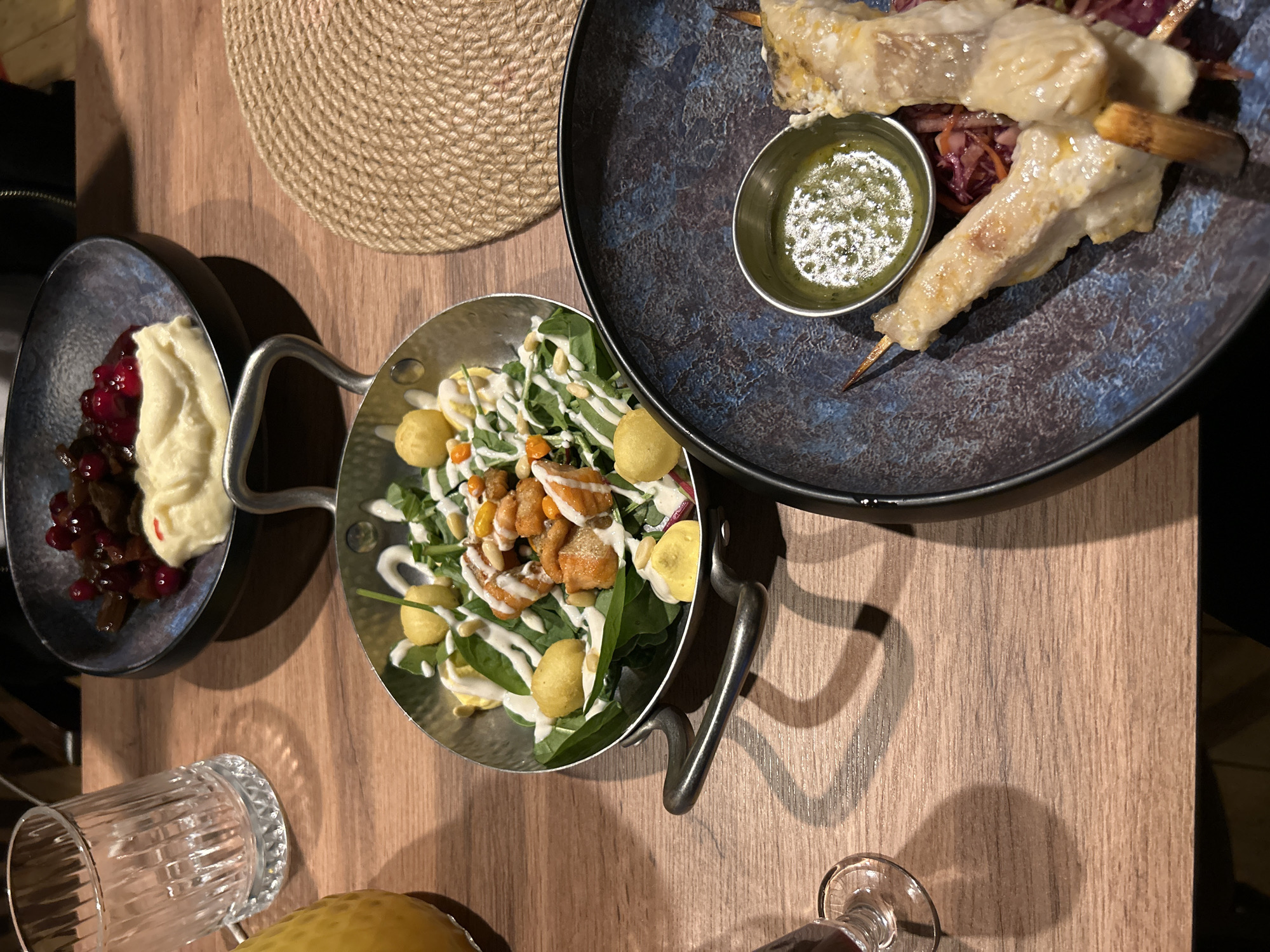 A table in a restaurant in Saint Petersburg, Russia, displays a meal. In the foreground, a dark blue bowl holds two skewers of white fish, accompanied by a small dish of green sauce and a bed of shredded red cabbage. To the left, a metal pan contains a salad with leafy greens, small yellow spheres, pieces of what appears to be fried fish or chicken, and pine nuts, all drizzled with a white sauce. A separate dark bowl in the upper background holds a portion of mashed potatoes topped with dark berries and what looks like pieces of meat. A clear glass with ribbed detailing sits to the left of the food, and a round, woven placemat is visible to the right. The setting appears to be indoors, with wooden table surfaces and shadows cast by objects on the table.