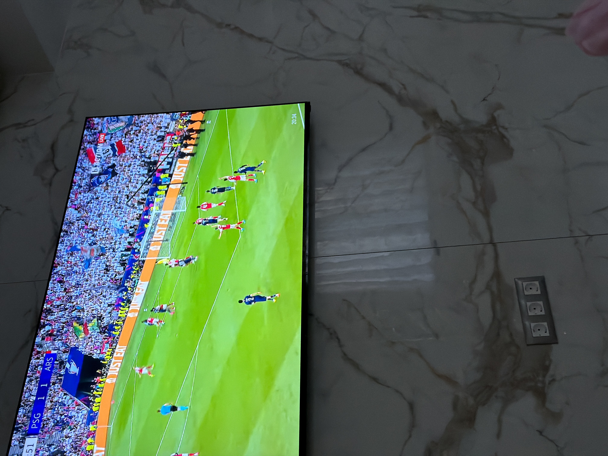 A television displays a soccer match, with the score PSG 1 - 1 ARS visible on the screen. The game shows a field view with many players present and a large crowd in the background. The time shown on the top right of the screen is 20:34. The TV is mounted on a wall with a marble-like pattern. To the right of the TV is an electrical outlet with three sockets. The image's location is uncertain, and there is no verifiable information to indicate that it is in Asenovgrad, Bulgaria. There are no people visible in the area of the photograph, other than players that can be seen on TV.