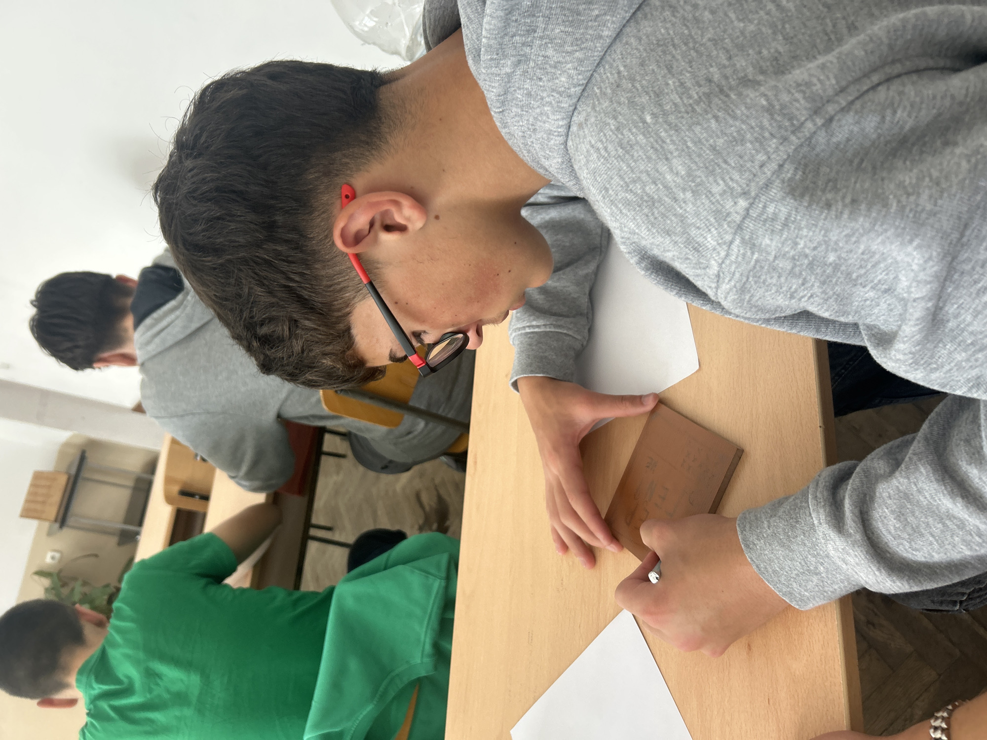 The image shows an indoor scene, likely a classroom, with multiple individuals. A young male wearing a grey hoodie and glasses is seated at a wooden desk, focused on tracing an object that appears to be a small plaque or block. He is using a pen and a piece of paper. Another person wearing a green shirt is visible at a nearby desk. In the background, there is another person wearing grey, partially visible. The location is presumed to be in Zlatograd, Bulgaria.
