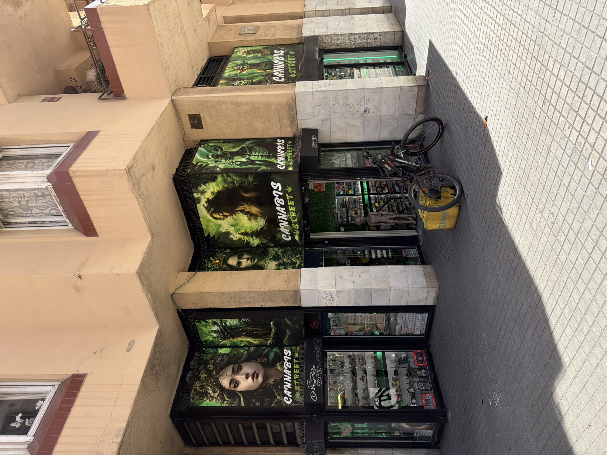 This image captures the exterior of a commercial establishment in Barcelona, Spain, likely a cannabis shop, given the prominent signage displaying "CANNABIS STREET" and imagery related to cannabis culture. The shop is situated at the corner of a building with a beige facade and brown window frames. Large, vibrant posters featuring stylized portraits and what appear to be artistic renderings of aliens and female figures adorned with cannabis leaves are plastered across the storefront.
The ground floor of the shop features display windows filled with various products, including what seem to be smoking accessories, lighters, and packaged goods. A bicycle, black with red accents, is parked leaning against a yellow delivery box labeled "Glovo" (a food delivery service). The pavement is tiled with dark gray square pavers, with a distinct shadow cast across it by the building. The overall impression is an urban street scene with a commercial focus on cannabis-related products. The building itself appears to be older, with some visible signs of wear and tear on the facade.