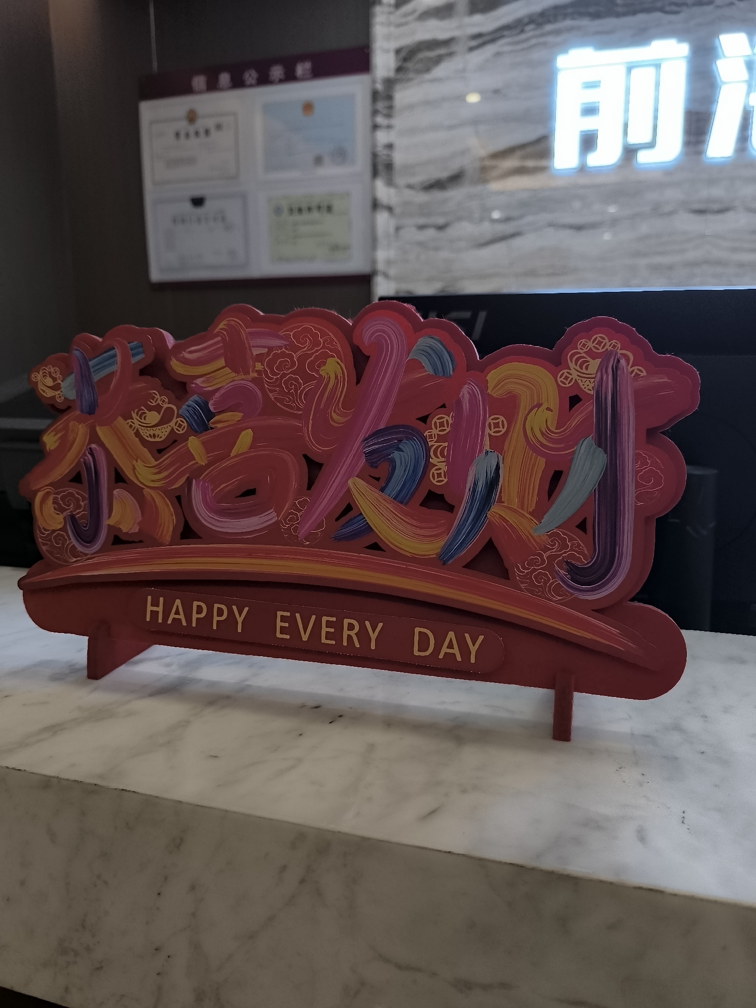 The image shows a red decorative piece with Chinese characters and the English phrase "HAPPY EVERY DAY" inscribed on a golden band. The Chinese characters appear to be festive and are rendered in a vibrant, brushstroke style with multiple colors. The decorative piece is placed on a white marble countertop. In the background, a blurred sign with Chinese characters and what appears to be a reception area or office can be seen. The sign in the background has the characters "前程" which translates to "future" or "prospects". Based on this context and the prevalence of such decor during Chinese New Year, this image was likely taken in Ürümqi, China, possibly in a business or hospitality setting, during a festive period.