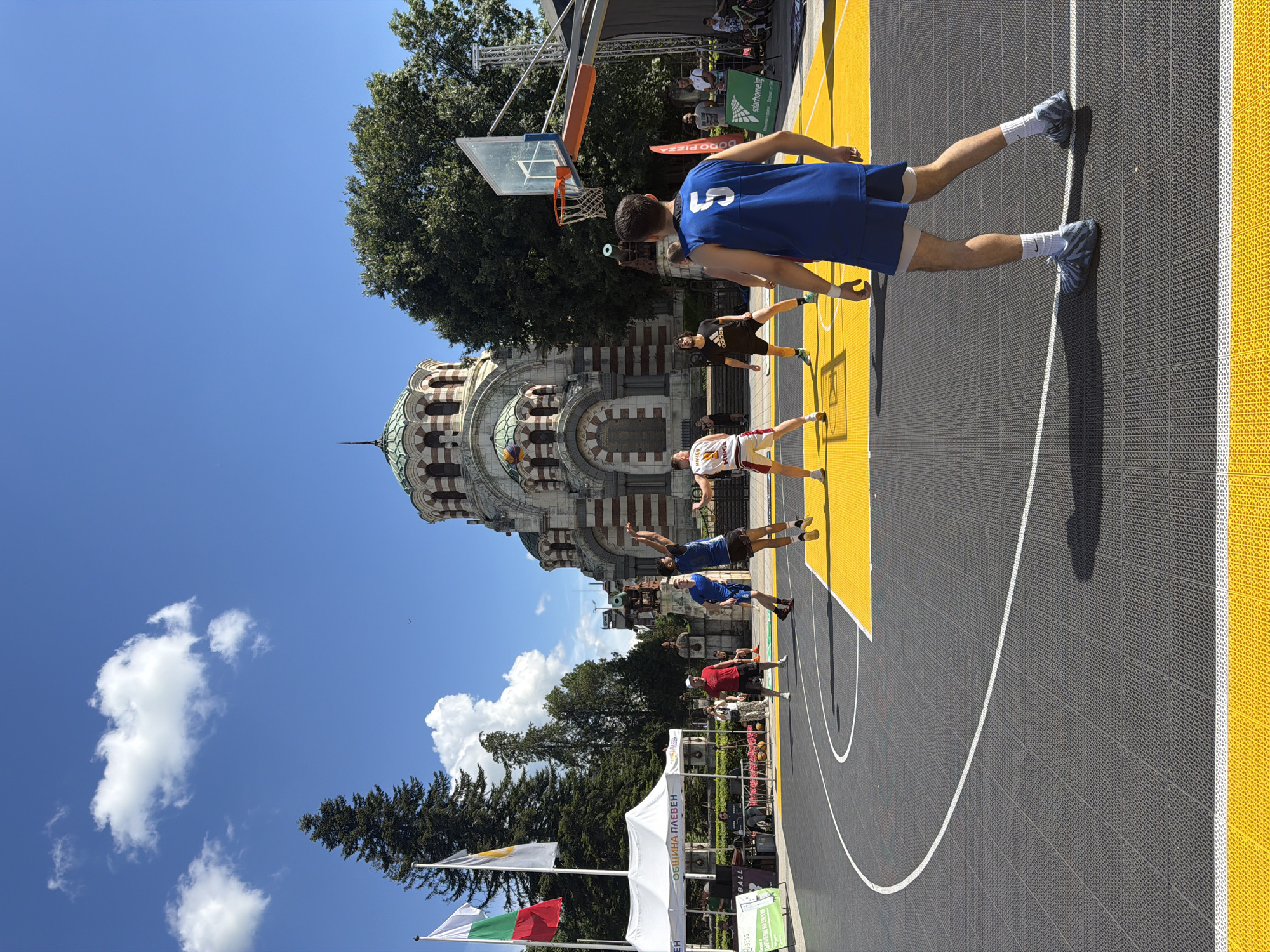 This image captures an outdoor basketball game taking place in front of the St. George the Conqueror Rotunda in Pleven, Bulgaria. The rotunda, a prominent landmark, features a distinctive dome and classical architectural elements, serving as a backdrop to the sports event. The basketball court is marked with yellow and gray tiles, with white lines indicating boundaries. Several individuals are actively participating in the game, including players in blue and white jerseys. One player, wearing a blue jersey with the number 5, is in the foreground, his back to the camera, appearing to be in a defensive stance. Other players are seen mid-action, with one jumping to shoot the ball. Spectators are visible in the background, some standing near tents and a stage setup. Bulgarian flags are displayed on poles to the left of the rotunda. The scene is set under a clear blue sky with scattered clouds, suggesting a sunny day. The overall atmosphere appears to be one of a community event or tournament.