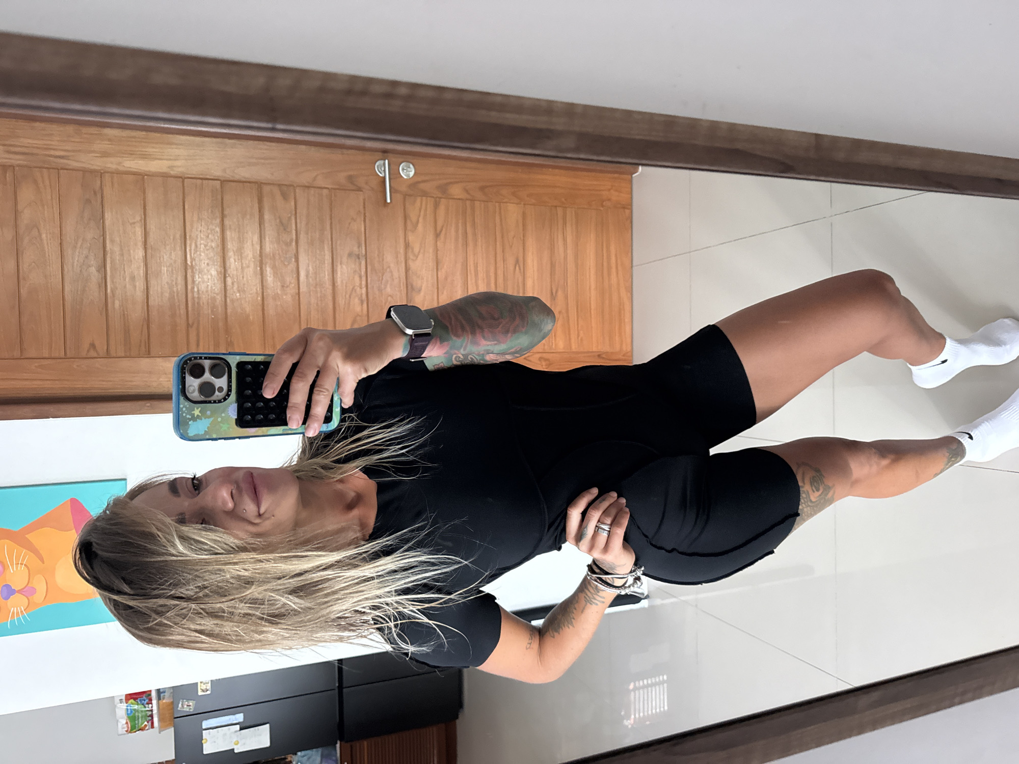 A woman with blonde hair, wearing a black athletic outfit and white socks, is taking a mirror selfie. She has tattoos on her arms and legs. In the background, a wooden door with vertical panels is visible, along with a tiled floor and a wall with a painting of an orange cat. The image appears to be taken indoors, possibly in a home or gym.