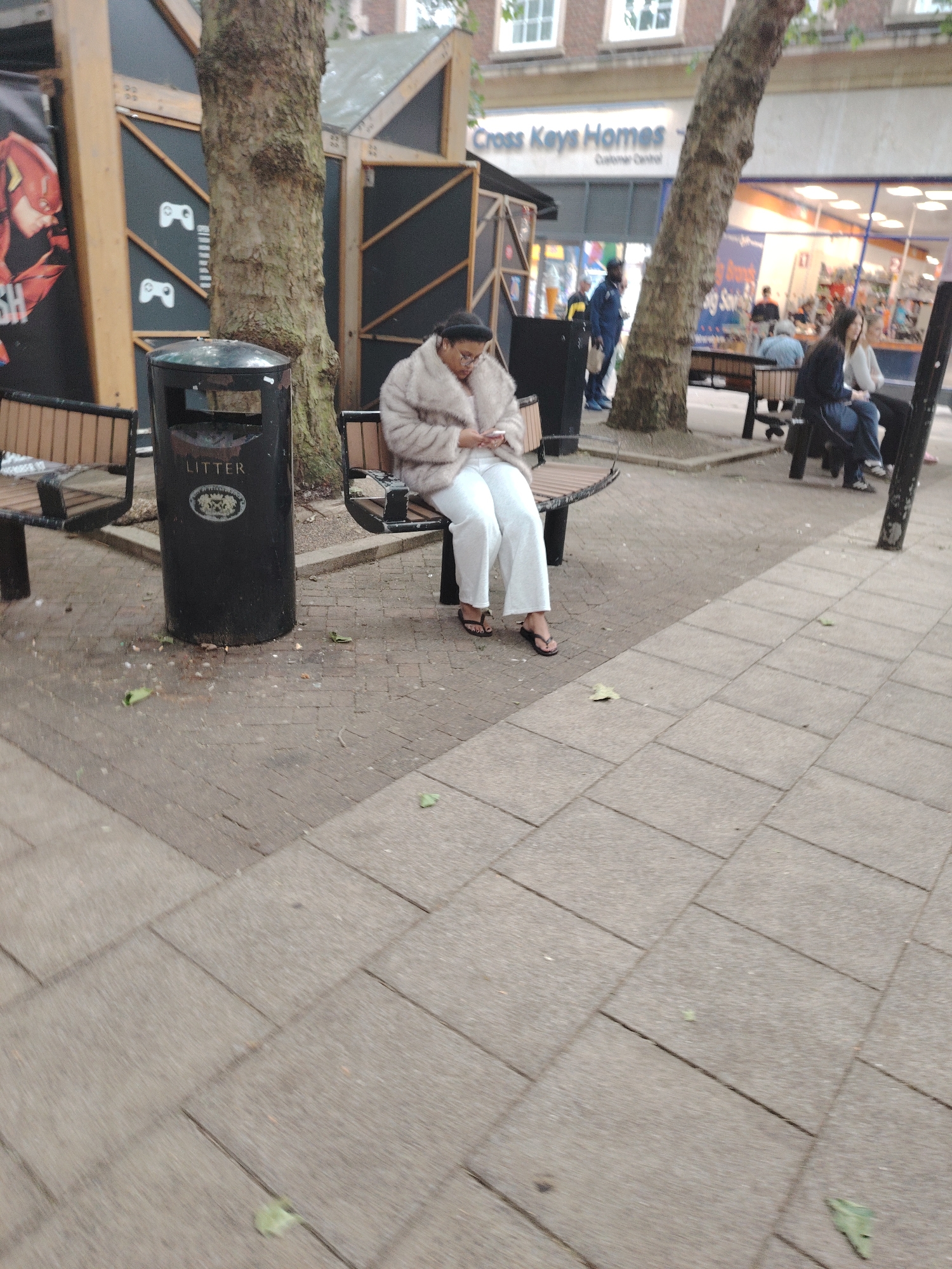 This image depicts an outdoor urban scene in Peterborough, United Kingdom, specifically a paved pedestrian area lined with commercial establishments. In the immediate foreground, a person, likely female, with dark hair and glasses, sits on a public bench. She wears a light-colored, possibly faux fur, jacket, white trousers, and black flip-flops, and is engrossed in a mobile phone held in her hands. Adjacent to her bench, a black cylindrical litter bin, embossed with the word "LITTER" and a metallic crest, stands on the paving. Scattered leaves and several cigarette butts are visible on the ground around the bin's base.
Behind the person on the bench, a mature tree trunk with rough bark rises. To the left, a structure displays a large poster featuring a red and yellow figure resembling The Flash, alongside dark panels adorned with white game controller icons. In the midground, another public bench is occupied by two individuals; one wears a dark top and blue jeans, looking towards the left.
The background features commercial buildings. A sign for "Cross Keys Homes" is prominently displayed on one building. To its right, a brightly lit shop interior reveals multiple indistinct figures. A person in a dark blue uniform, carrying a yellow object on their back, is visible walking from left to right near the "Cross Keys Homes" building. The ground is uniformly covered with rectangular grey and beige paving slabs, interspersed with occasional leaves.