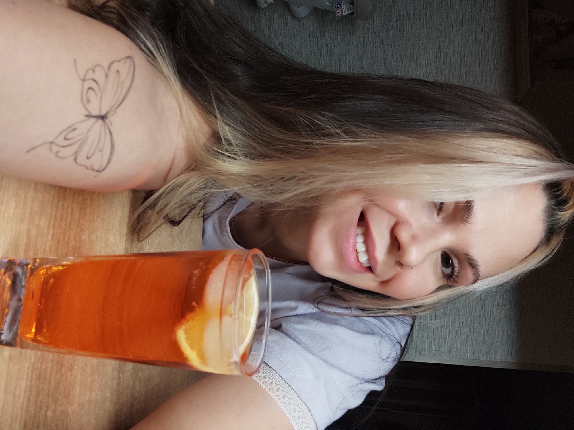 A young woman is smiling at the camera. She has dark hair with blonde highlights and is wearing a light-colored t-shirt with white lace trim. A glass of orange liquid with a lemon slice is next to her. A butterfly tattoo is visible on her left shoulder. The background is a textured wall and a wooden surface. The image appears to have been taken indoors. The specific location in the city of Chaikovsky, Russia is unknown.
