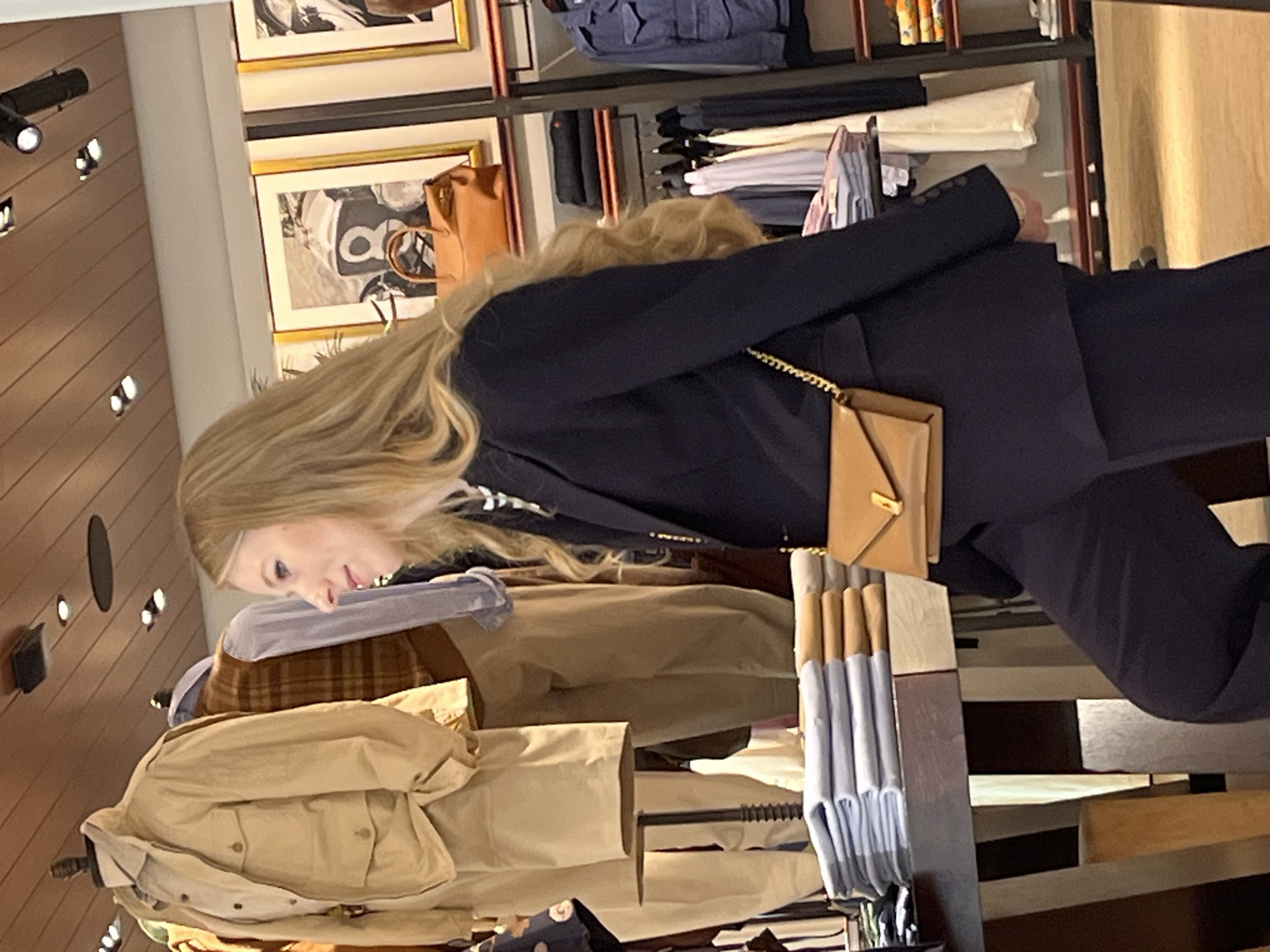 The image captures a candid indoor scene, likely within a retail clothing store in Barcelona, Spain. A blonde woman, viewed from the chest up and slightly from the side, is the central subject. She wears a dark blue blazer and matching trousers, and a small, rectangular light brown leather shoulder bag with a gold chain strap is visible across her body. Her face is in profile, looking downwards and to her left, with a neutral, contemplative expression. Her long, blonde hair flows over her shoulders and down her back.
The background reveals typical retail displays. To the woman's right, there are racks of clothing, including folded light-colored shirts (some blue, some white) and a beige jacket. Above these, shelves hold more folded garments, including what appear to be denim shirts or jackets. On the wall, several framed pictures with golden frames are hung, depicting what seem to be vintage-style black and white illustrations or photographs, possibly related to fashion or lifestyle, though the details are unclear.
The ceiling features recessed spotlights, illuminating the space. A light-colored wooden slatted wall or ceiling design is also visible in the upper left. Another clothing item, a plaid-patterned garment, can be partially seen behind the woman. The overall impression is that of an upscale and well-lit boutique. There is no visible text in the image.