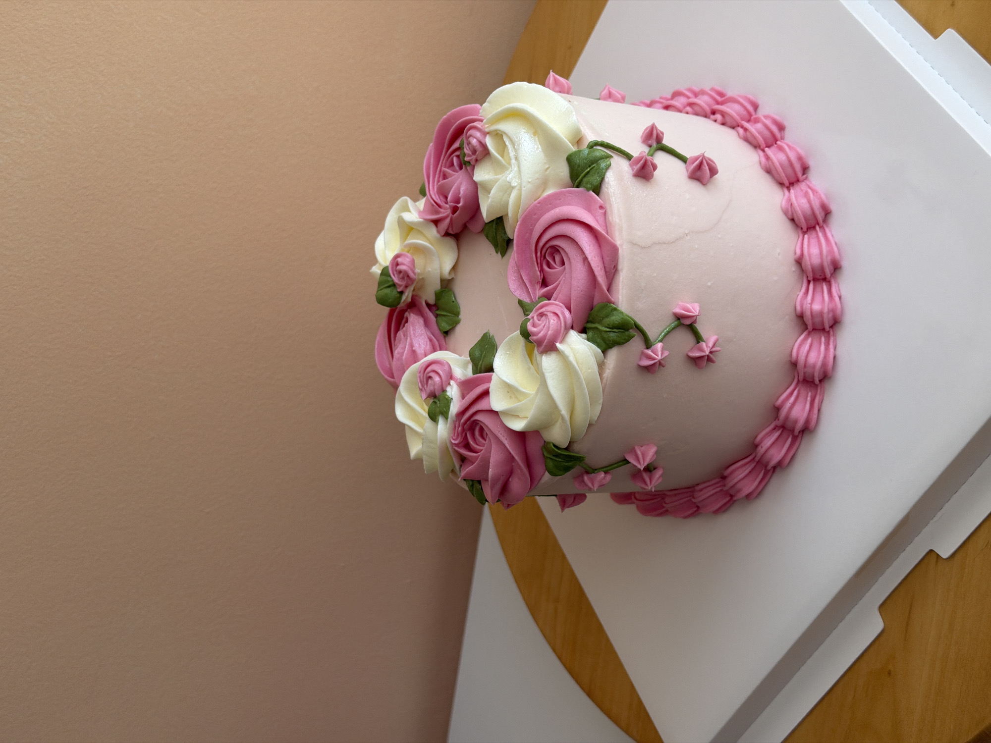 A close-up, eye-level shot shows a pink frosted cake with decorative piping. The cake is adorned with alternating pink and cream-colored buttercream roses and green leaves arranged in a circular pattern on top. A delicate vine with small pink buds and leaves trails down the side of the cake. A pink piped border encircles the base of the cake. The cake rests on a white cake board, which is placed on a light-colored surface with a wooden element visible at the bottom left. The background is a plain, textured peach-colored wall. The image does not contain any identifiable landmarks or clues to suggest a location in Tel-Aviv, Israel.