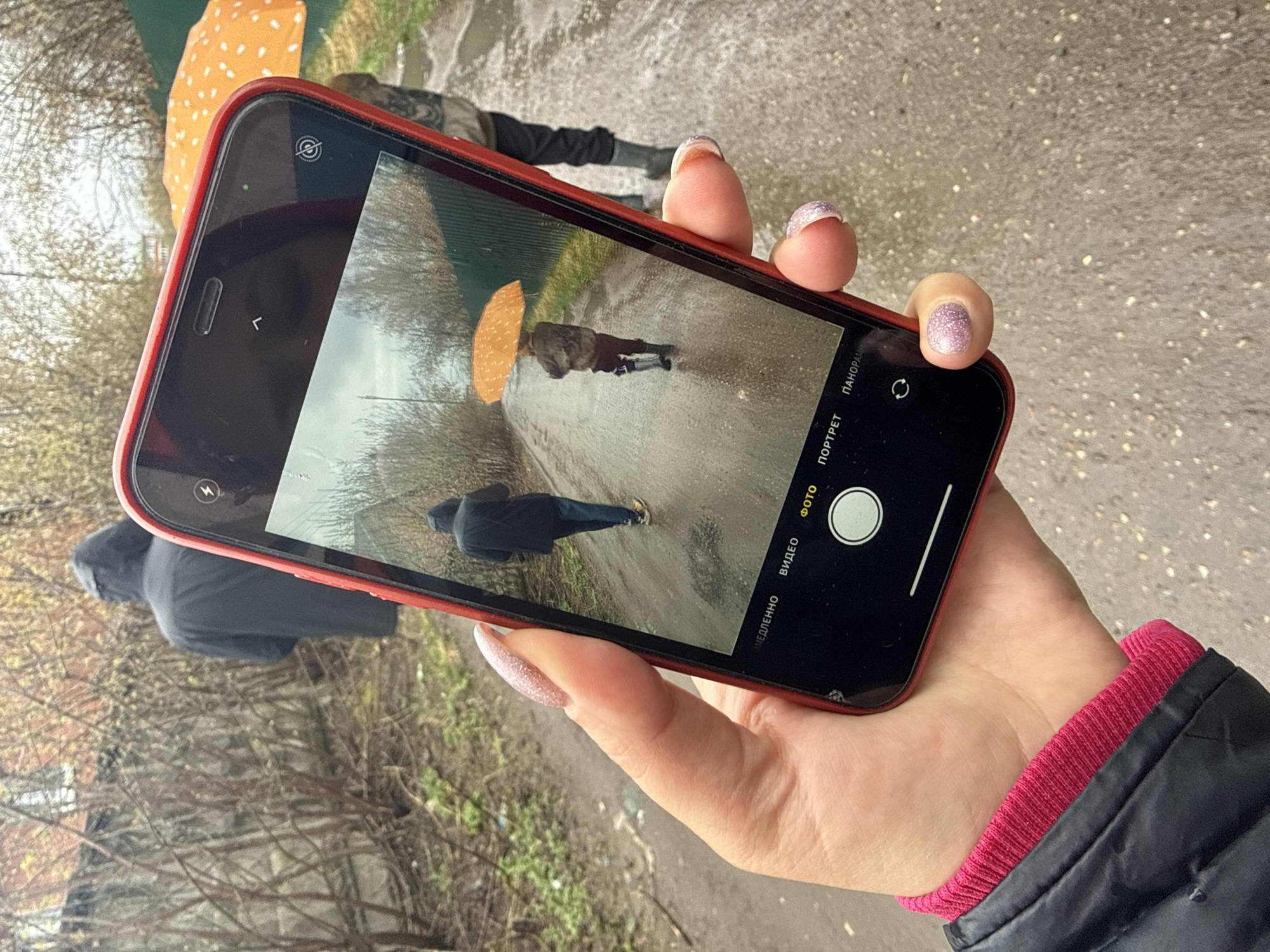 A person holds a red iPhone, displaying a photo of a rainy scene with two people walking on a path. One person in the photo holds an orange umbrella. The photo appears to be taken in Dmitrov, Russia, based on the environment. The ground is wet, indicating rain. The person holding the iPhone has manicured fingernails with glittery polish and is wearing a black jacket with a pink cuff. Another person, in a black hooded jacket, stands nearby, partially visible in the background. The area consists of vegetation and a paved path.