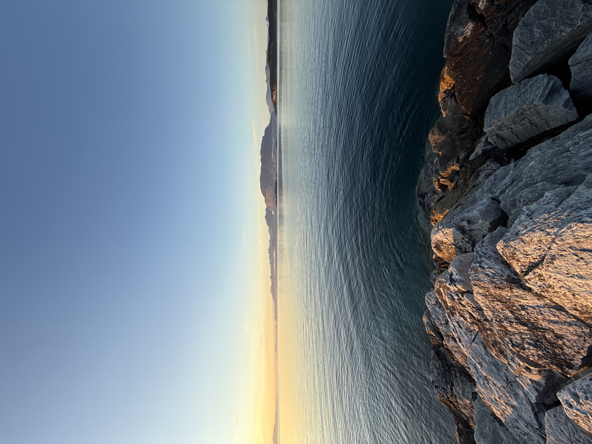 This image depicts a serene coastal scene at dawn or dusk in Harstad, Norway. In the foreground, a pile of rugged, grey rocks, some tinged with warm golden light, form the edge of the land. Beyond the rocks, a vast expanse of calm, deep blue water stretches towards the horizon. The water is characterized by gentle ripples and reflects the soft colors of the sky. In the distance, a chain of silhouetted mountains with softly rounded peaks emerges from the water, partially shrouded in mist or low-lying clouds. The sky above is a gradient of soft blues, transitioning to a pale yellow and orange hue near the horizon, indicating the sun's low position. The overall atmosphere is peaceful and tranquil.