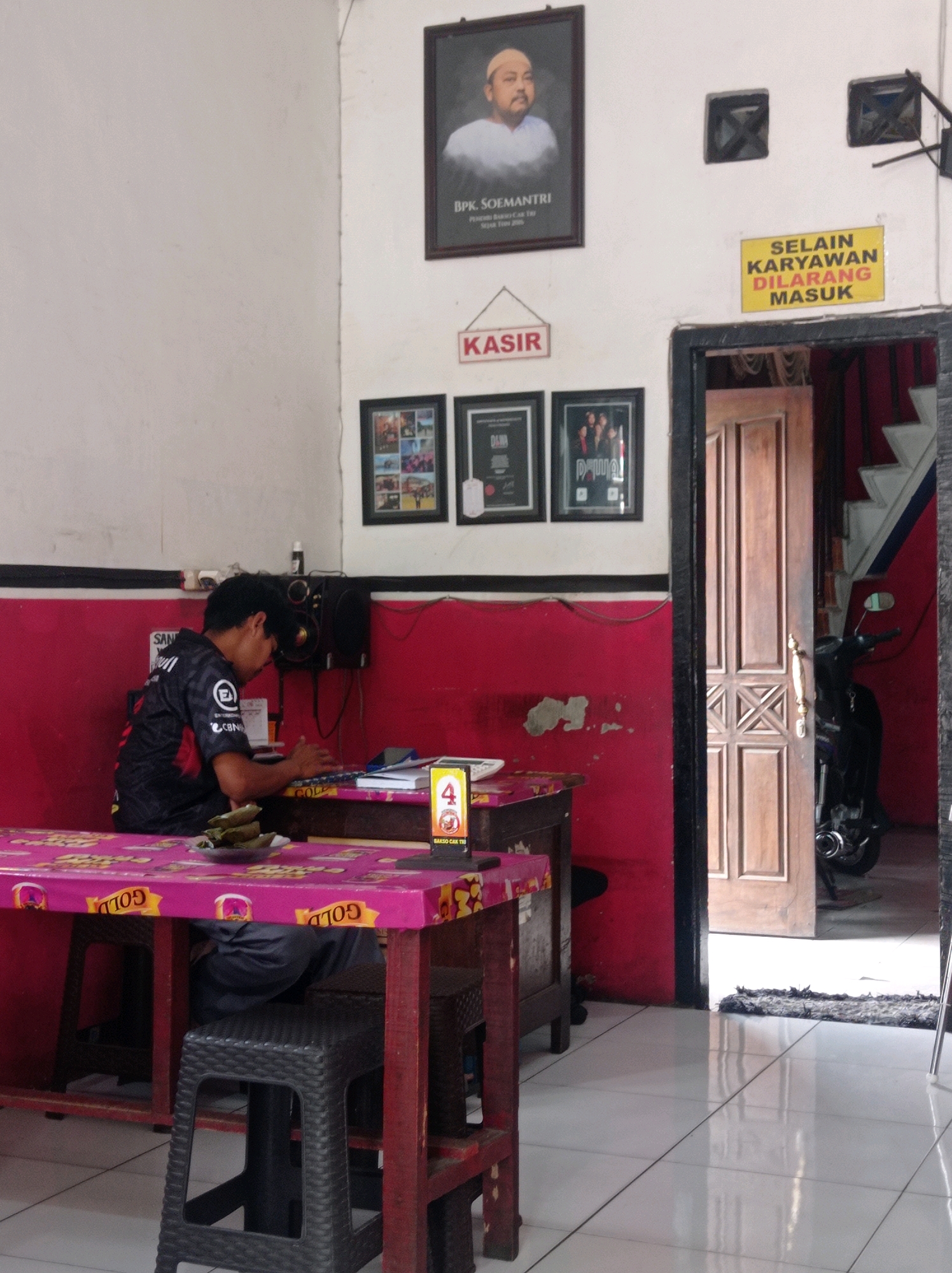 The image depicts the interior of a food establishment, likely a restaurant or warung, in Kedok, Indonesia. A young man, wearing a black and gray patterned shirt with the logo "CBN" visible, is seated at a table and appears to be working on a laptop. The table is covered with a pink tablecloth featuring a repeating pattern. On the table, there are some food items wrapped in leaves and a plate. A speaker system is mounted on the wall behind him.
Above the man, on the wall, are several framed items. A large portrait of a man in a white shirt is prominently displayed, with text below it reading "BPK. SOEMANTRI" and other smaller text. To the right of this portrait, a yellow sign with black text reads "SELALAIN KARYAWAN DILARANG MASUK," which translates to "ONLY EMPLOYEES ARE ALLOWED TO ENTER." Below the portrait and the yellow sign, a smaller red sign reads "KASIR," indicating a cashier area.
To the right of the frame, a wooden door stands open, revealing a glimpse of stairs and a motorcycle parked inside. The floor is tiled with white square tiles. The overall environment suggests a functional and moderately decorated interior of a local business.