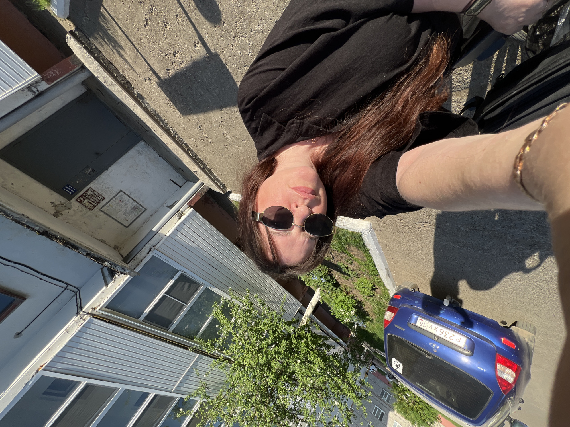 A rotated upward-angle photograph depicts a person with long brown hair, wearing dark sunglasses and a black shirt, positioned near a building and a blue car. The individual is holding the camera arm outstretched. The building has grey metal siding and visible windows. Adjacent to the building, a tree with white blossoms is partially visible. A blue car is parked near the building with the license plate P236XY 18. The surrounding environment appears to be an urban area, and is potentially located in Noviy, Russia.
