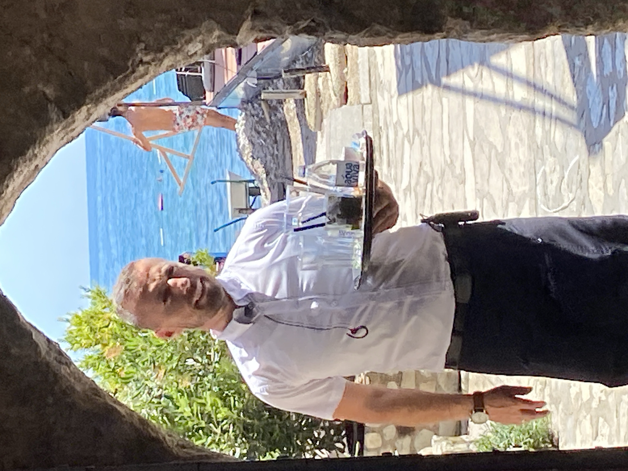 The image portrays a bright, sunlit coastal scene in Budva, Montenegro, viewed through a dark, rough-hewn stone archway. A Caucasian male, identified as a waiter, stands in the foreground, centrally framed by the archway. He has short, light-colored hair and stubble, displaying a slight smile. He wears a short-sleeved white button-up shirt, a dark apron/waistcoat, and a brown-strapped watch on his left wrist. A dark earphone with a red tip and cord is visible around his neck. He carries a dark rectangular tray in his left hand, holding multiple clear drinking glasses, a small plastic bottle of "Aqua Viva" mineral water, and a small white cup. A dark object is clipped to his right hip. The ground beneath him is paved with light-colored, irregularly shaped stones.
In the middle ground, beyond the waiter, a clear blue sea stretches to the horizon under a bright sky. Lush green foliage is visible on the left side of the archway opening. To the right, a shirtless Caucasian male with dark hair, wearing patterned swim trunks, stands near the water's edge, holding a light-colored, A-frame or triangular structure. In the distant water, a pink inflatable float and a swimmer are visible. The shoreline includes a light-colored wheelbarrow on the left and a glass barrier/railing on the right, separating a paved area from the rocky beach. A distinct triangular shadow, consistent with bright daylight, is cast on the paved ground to the right of the waiter.