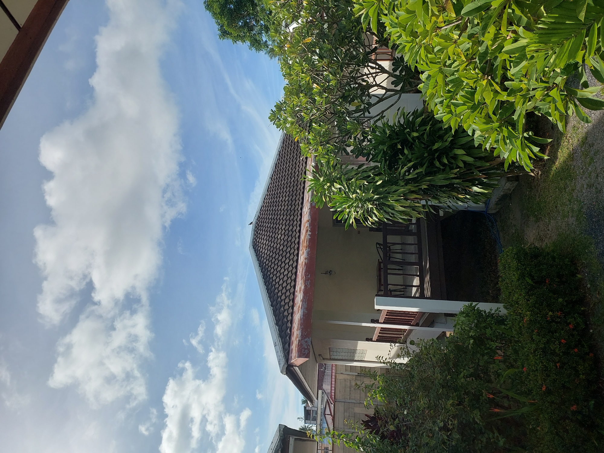 The image shows a bungalow in Baan Mae Nam, Thailand, with a tiled roof and a wooden veranda. Lush green foliage surrounds the bungalow, with a few small orange flowers visible. A table and chairs are present on the veranda. The sky is partly cloudy, with a few wispy clouds against a blue background. The ground appears to be a mix of gravel and grass. A bird is perched on the roof of the bungalow.
