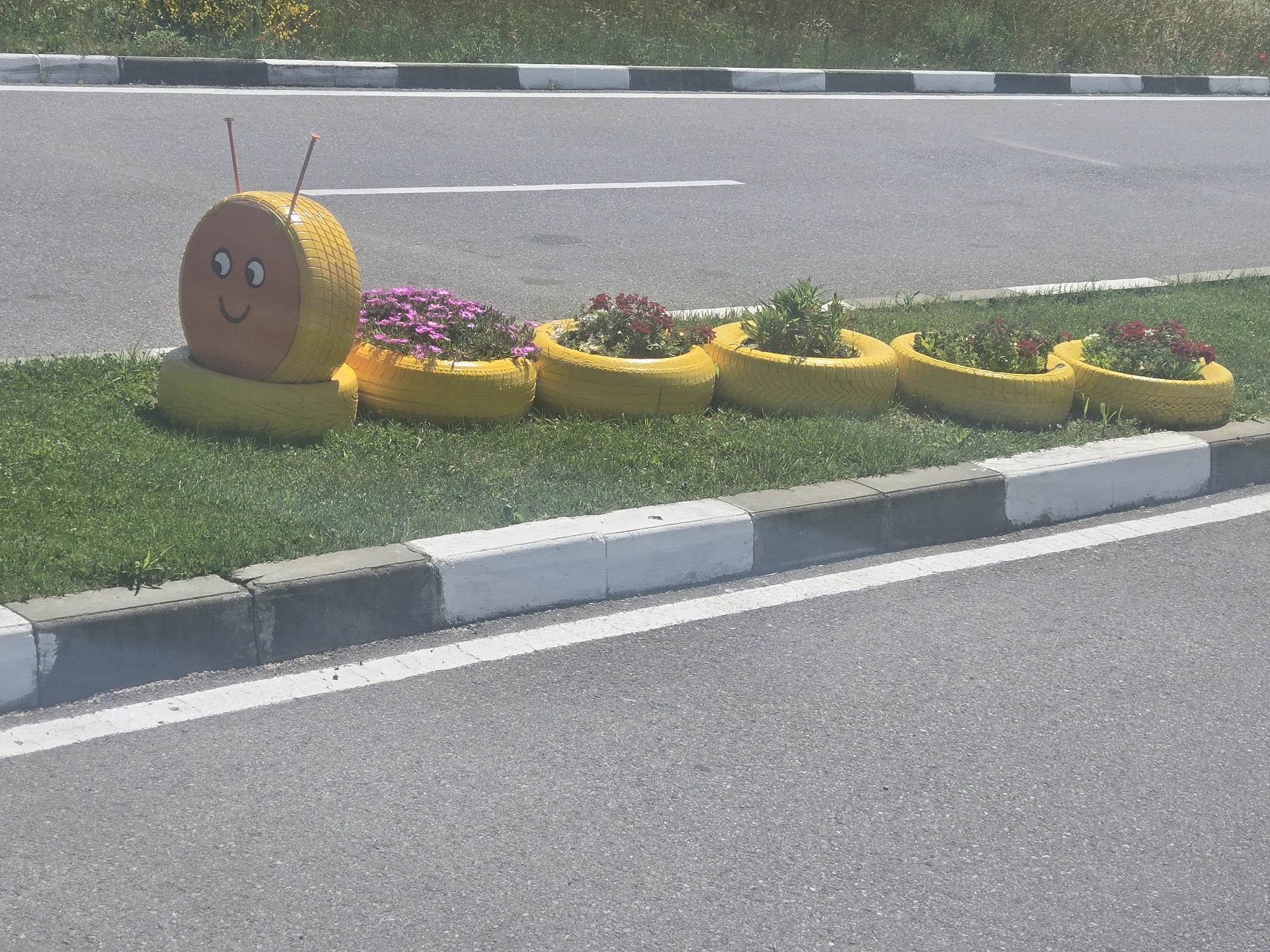 The image shows a roadside decoration in the form of a caterpillar made from yellow painted tires, serving as flower planters. The caterpillar's head, positioned to the left, has a painted face with eyes and a smile, and antennae made of thin rods. The body consists of five tires filled with various flowers and greenery. The caterpillar is placed on a strip of grass between the road and the curb. The road is made of asphalt with a white line painted along its edge. The location appears to be an urban or suburban area, in the city of Unknown, Unknown, possibly near a residential or commercial zone. The time of day is likely daytime due to the natural lighting. There are no people visible in the scene.