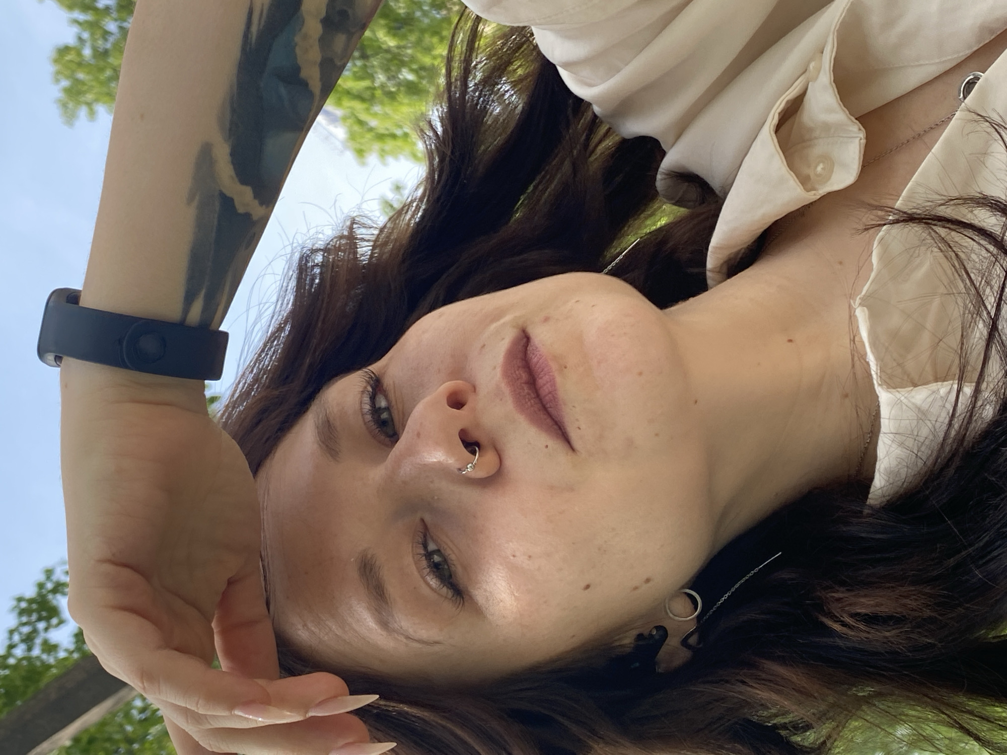 A person is viewed from a low angle against a backdrop of trees and a blue sky. The person is wearing a light-colored button-down shirt and has brown hair. A tattoo is visible on their left arm. They have a nose ring and are wearing a black wristband. Their hand is raised near their face. The location appears to be outdoors in a wooded area. No information is available to confirm the location as Chaikovsky, Russia.