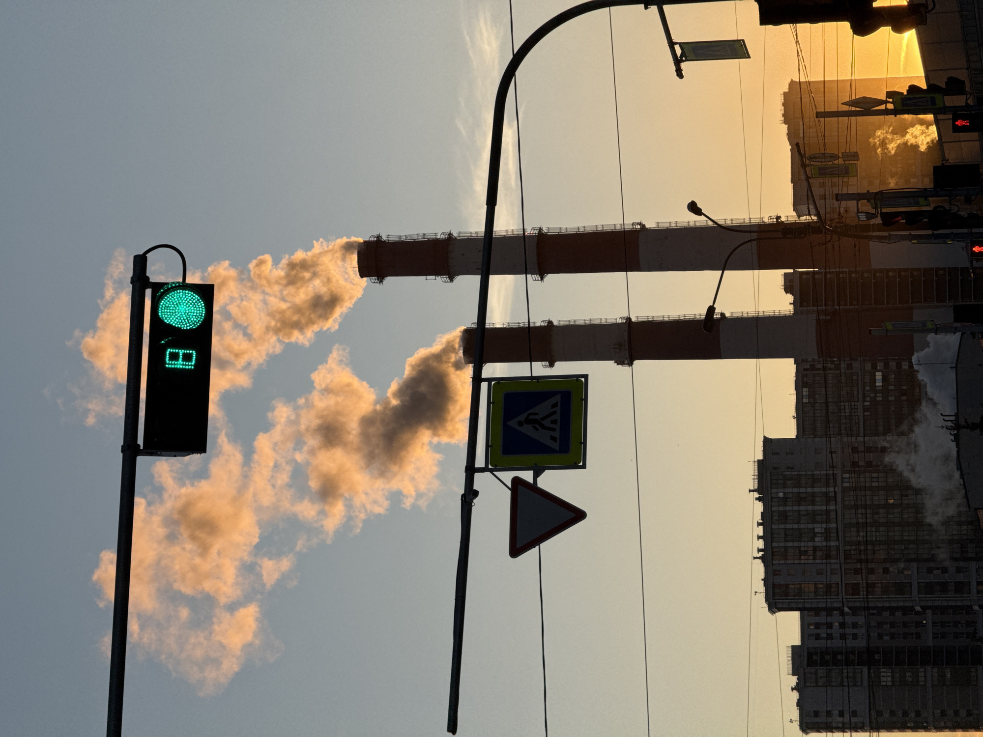 The image depicts an urban landscape at sunset, likely in Saint Petersburg, Russia, given the architectural style and industrial elements. In the foreground, a green traffic light stands illuminated, contrasting with the fading daylight. Adjacent to it is a pole with a pedestrian crossing sign and a triangular warning sign. In the background, two tall industrial chimneys emit plumes of smoke into the sky, tinted orange by the setting sun. Several apartment buildings are also visible, their silhouettes outlined against the sky. Power lines are strung across the sky, adding to the urban character of the scene. The overall impression is one of a city coexisting with its industrial infrastructure, framed by the warm light of the sunset.