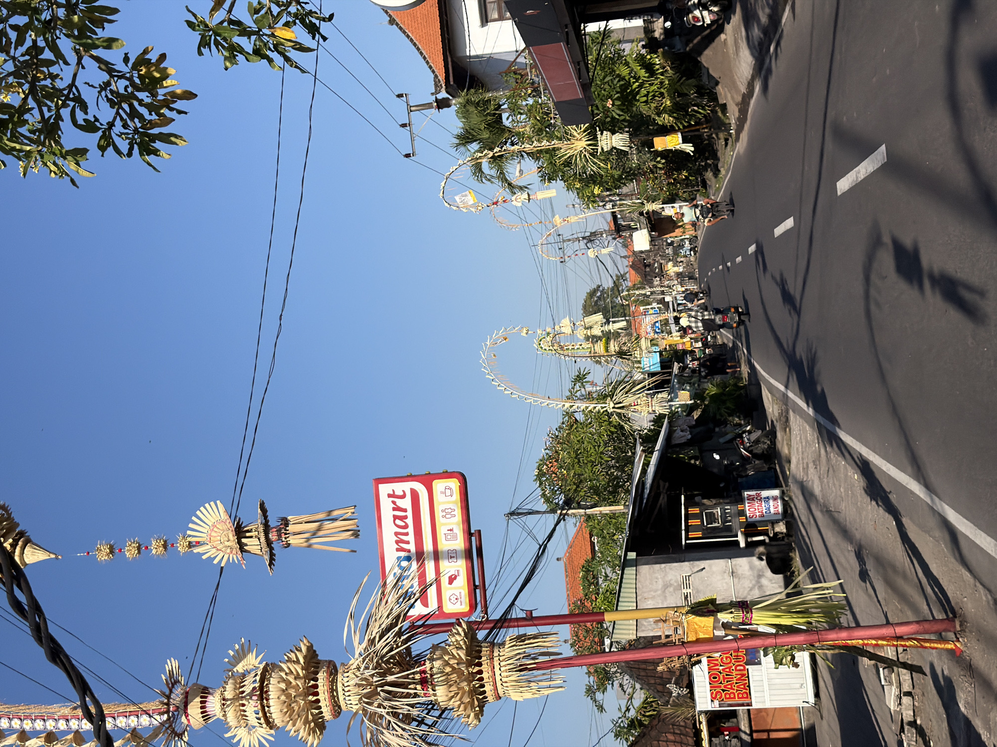 The image depicts a street scene in Mengwi, Indonesia, likely during a festive period, given the numerous ornate, palm-leaf decorations adorning lampposts and utility wires. A prominent red and white sign for "Indomaret" is visible on the left, indicating a convenience store. The street itself is paved and appears to be a public thoroughfare with moderate traffic, including motorcycles and what appear to be a few cars further down the road. Several individuals are seen on motorcycles, navigating the street. Buildings lining the street showcase typical Balinese architecture, with tiled roofs. Vegetation, including trees and bushes, is abundant. The sky is clear and blue, suggesting a sunny day. The overall atmosphere suggests a vibrant local street, possibly in a semi-urban or suburban area.