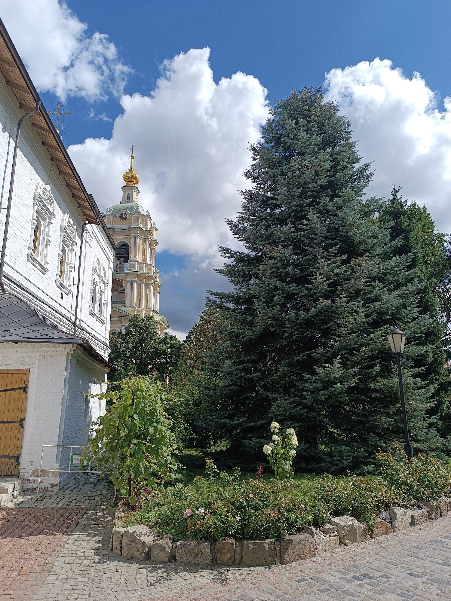The image depicts the bell tower and adjacent building of the Novodevichy Convent in Moscow, Russia. The bell tower, with its green dome and golden cross, stands tall against a backdrop of a bright blue sky with fluffy white clouds. In the foreground, a white brick building with decorative window frames is partially visible. Lush green trees, including a large blue spruce, dominate the right side of the frame. A path paved with cobblestones leads towards the buildings. A street lamp stands amidst the greenery. Several bushes and flowering plants, some with pink roses, are arranged in a garden bed bordered by large stones. A grapevine is also visible near the white building. The scene conveys a sense of peacefulness and historical significance.