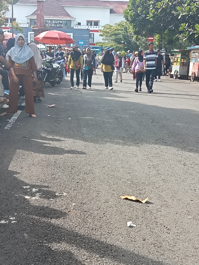 The image captures a vibrant daytime street scene in Singaparna, Indonesia, likely during a public event or gathering. Numerous people are present, with many walking along a paved road and others observing from the side. A significant number of women are seen wearing hijabs, indicating a Muslim-majority population.
In the foreground and midground, groups of individuals are walking towards the viewer and further down the street. Their attire is casual, suggesting a leisurely activity. On the far left, several people are seated or standing near parked motorcycles, partially obscured by a red umbrella. To the right, a line of street food carts is visible, indicating local commerce or a market setup.
The background features a large building with a red roof, which has visible text that reads "KANTOR BUPATI" and partially "MALAYA," identifying it as the Regent's Office, likely referring to Tasikmalaya Regency, where Singaparna is located. A blue sign and a red no-entry sign are also discernible near the building.
The scene is brightly lit by natural sunlight, casting distinct shadows on the asphalt road, suggesting a clear day in the morning or early afternoon. Scattered litter, including a dried leaf and small pieces of white paper, is visible on the road surface. The overall atmosphere conveys a sense of community activity and movement.