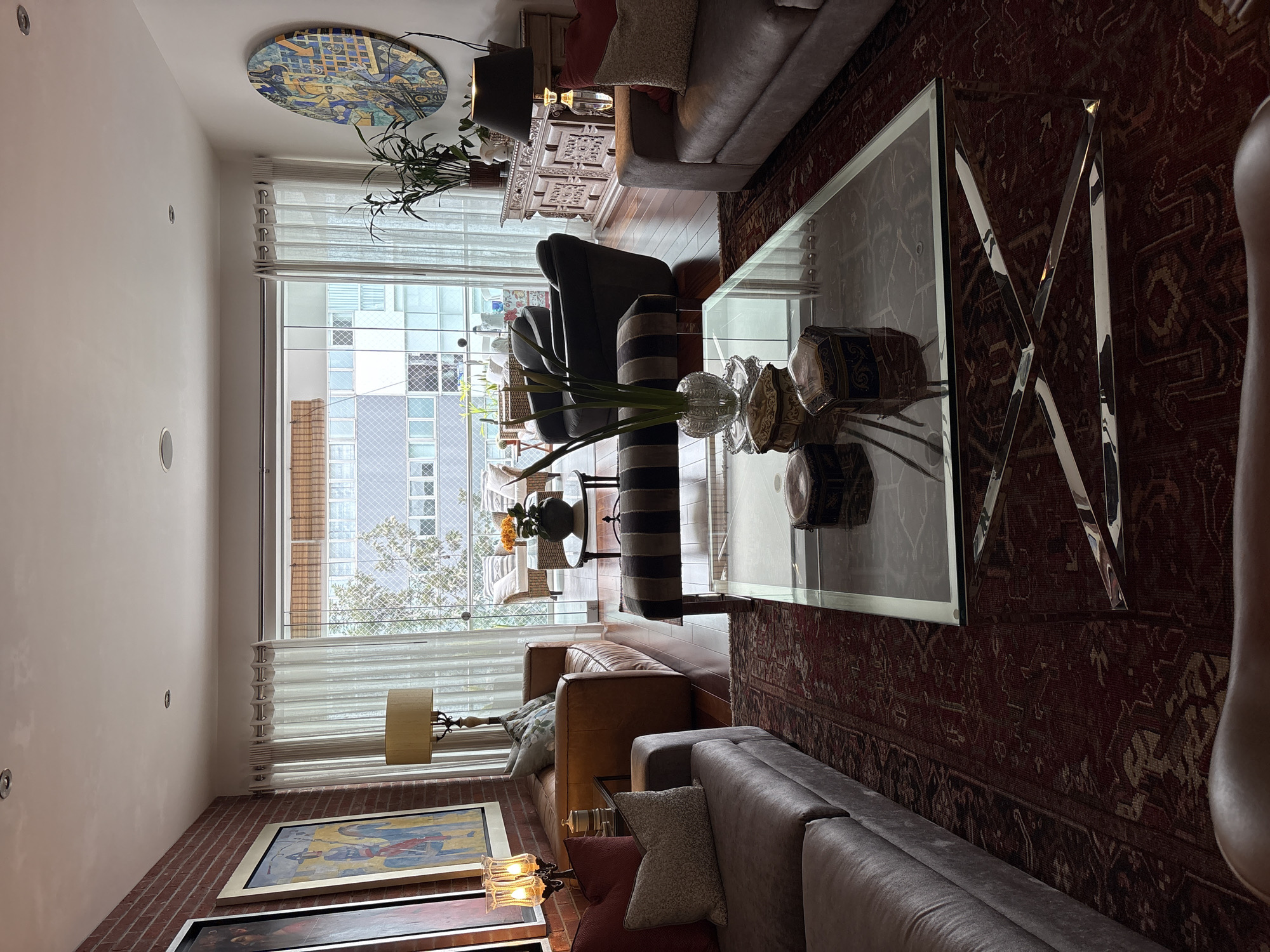 The image depicts a well-appointed living room in a city apartment in San Isidro, Peru. The room is furnished with two large, plush sofas, one a dark grey and the other a lighter grey, positioned to face a glass and chrome coffee table. On the coffee table are several decorative boxes and a vase with greenery. A striped bench sits in front of the coffee table. To the left of the sofas, a brown leather sofa is visible, accompanied by a floor lamp with a cream-colored shade and a brick wall as a backdrop. The room is adorned with two large framed paintings on the brick wall.
The living room opens to a balcony or patio area through large sliding glass doors, where outdoor furniture, including chairs and a small table with flowers, is set up. Beyond the balcony, the urban landscape of San Isidro is visible, with apartment buildings and trees. A large, circular, colorful artwork is displayed on the white wall to the right of the sliding doors, near a wooden credenza topped with a lamp, plants, and decorative objects. The flooring throughout the apartment appears to be polished dark wood, and a large, intricately patterned rug in shades of red and brown covers a significant portion of the living room floor. The lighting in the room is a mix of natural light from the balcony and artificial light from lamps. No people are present in the scene.