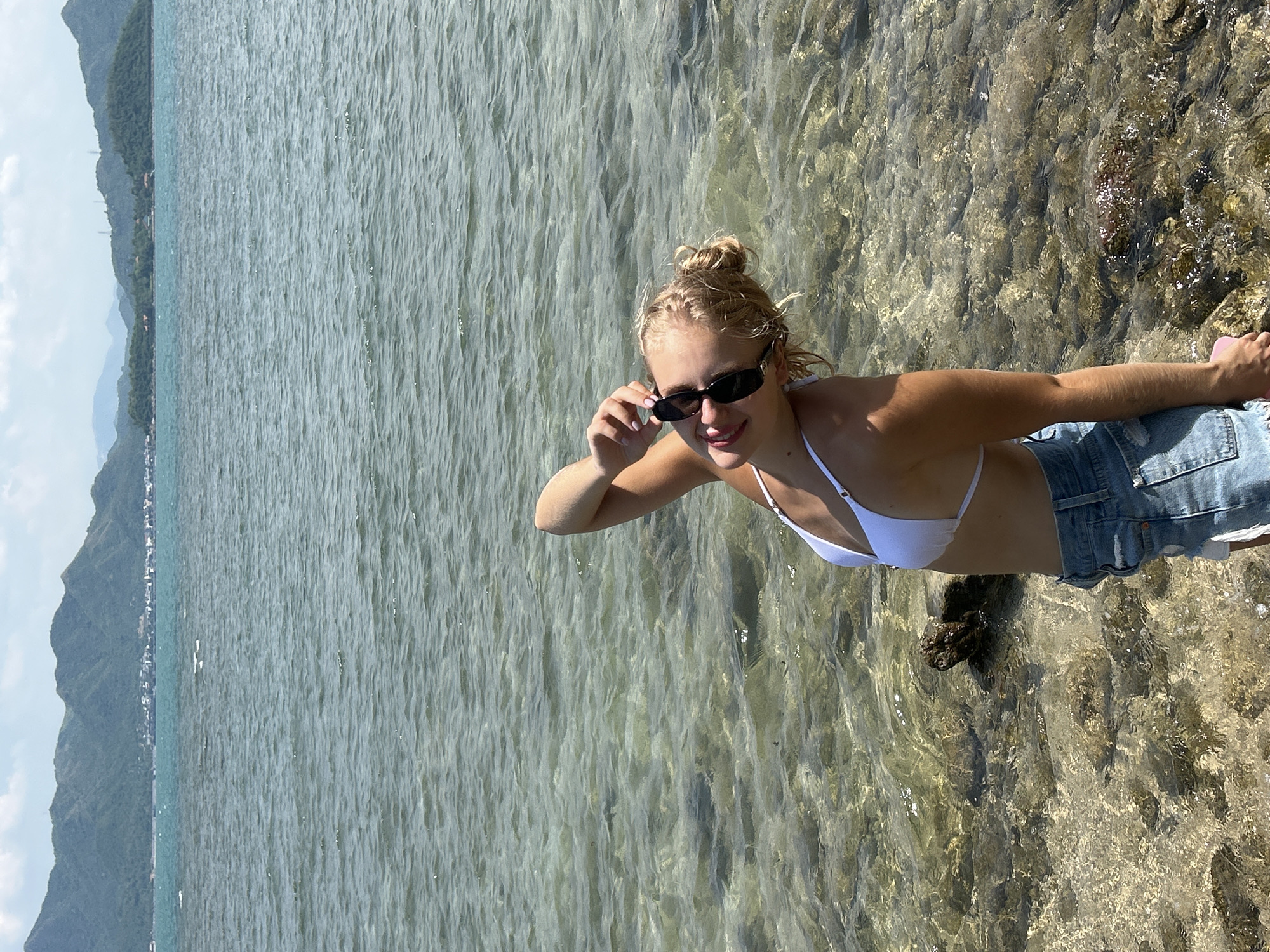 A young woman stands at the edge of clear, shallow water on a rocky shore, with a distant view of mountains and a coastal town. She is wearing a white bikini top and denim shorts, and sunglasses. Her hair is pulled up in a bun, and she is tilting her head back and touching her sunglasses with her right hand. The water is calm, with sunlight reflecting off the surface and illuminating the rocky seabed. In the background, lush green mountains rise from the horizon, with a few buildings visible in the distance, suggesting a populated coastal area. The sky is partly cloudy, casting soft shadows. The overall scene depicts a peaceful day at the beach in a scenic coastal location.