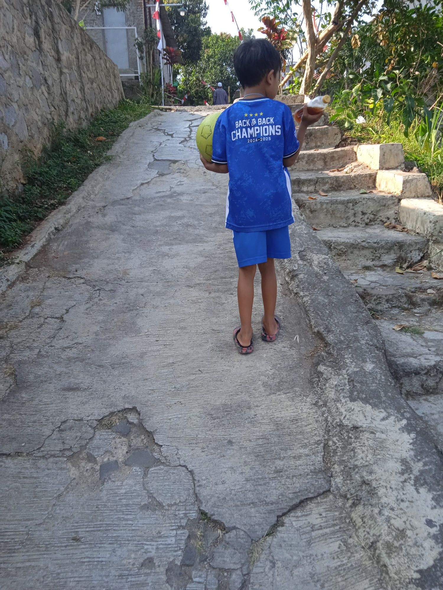 A young boy, facing away from the camera, stands on a cracked concrete pathway that slopes upwards. He is wearing a blue t-shirt with white text and graphics that read "BACK TO BACK CHAMPIONS 2024-2025" and blue shorts. He holds a yellow ball in his left arm and a bottle of drink in his right hand. To the right of the pathway, there are stone steps leading upwards, partially overgrown with vegetation. A stone wall borders the left side of the pathway, with green plants growing along its base. In the background, trees and a building can be seen. The scene appears to be in a residential area of Cileunyi, Indonesia, likely during daylight hours.