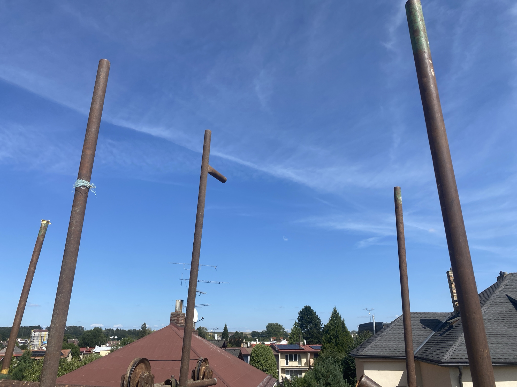 This is a low-angle, full outdoor shot of a residential area under a bright blue sky with thin, wispy clouds. The foreground is dominated by four tall, rusty metal poles, some of which are partially obscured by the edges of the frame. One pole on the far left has a light blue string tied around it. Another pole in the middle has a horizontal piece attached, resembling a cross or a T-junction. The middle ground reveals a cluster of houses with reddish-brown and dark grey pitched roofs, interspersed with trees and a few antennas on rooftops. The furthest background shows a treeline and more distant buildings. The scene appears to be in Polička, Czechia, suggesting a view from a rooftop or elevated position overlooking a neighborhood. There are no people visible in the image.