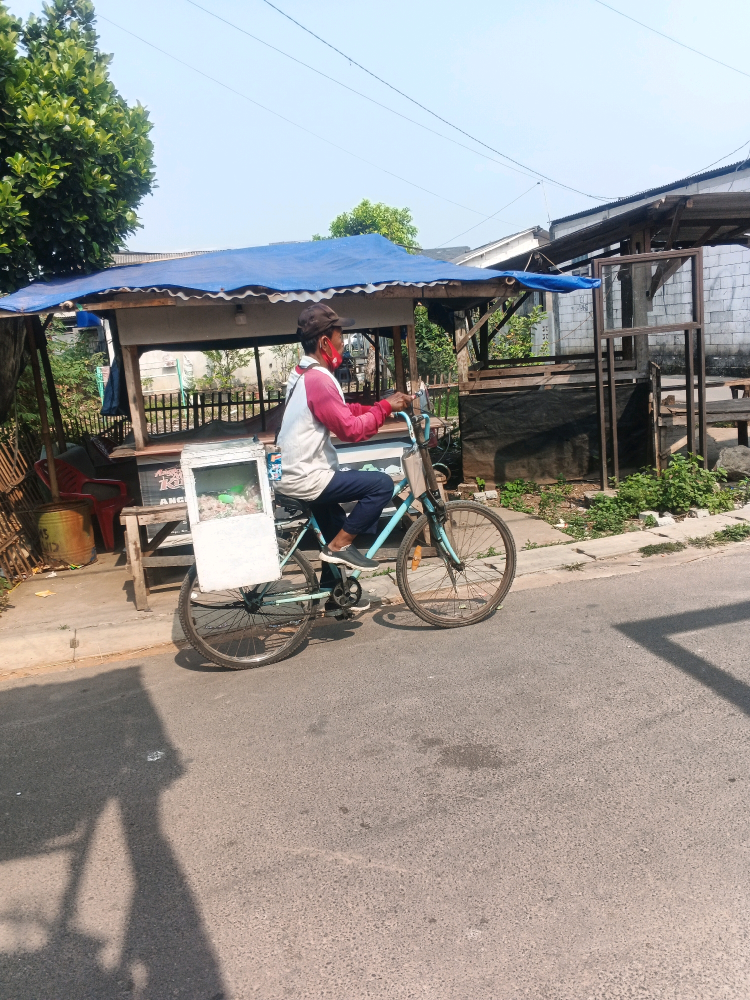 A man wearing a maroon baseball cap, a white long-sleeved shirt with maroon sleeves, and dark pants rides a light blue bicycle down a paved road in Kadu, Indonesia. He is wearing a red face mask. The bicycle has a white box attached to the rear, which appears to be a food or drink cooler. The man is pedaling the bicycle, which is in motion.
The background shows a street-side market stall with a blue tarp roof supported by wooden beams. A red plastic chair is visible under the stall, and a large, weathered metal barrel sits next to it. Behind the stall, there are more wooden structures and a white brick wall. Green foliage is growing around the market. The sky is clear and blue, indicating a sunny day. The road surface is asphalt, with a lighter-colored concrete curb on the left side of the image. Shadows are cast on the road from the man and bicycle, as well as from unseen objects.
