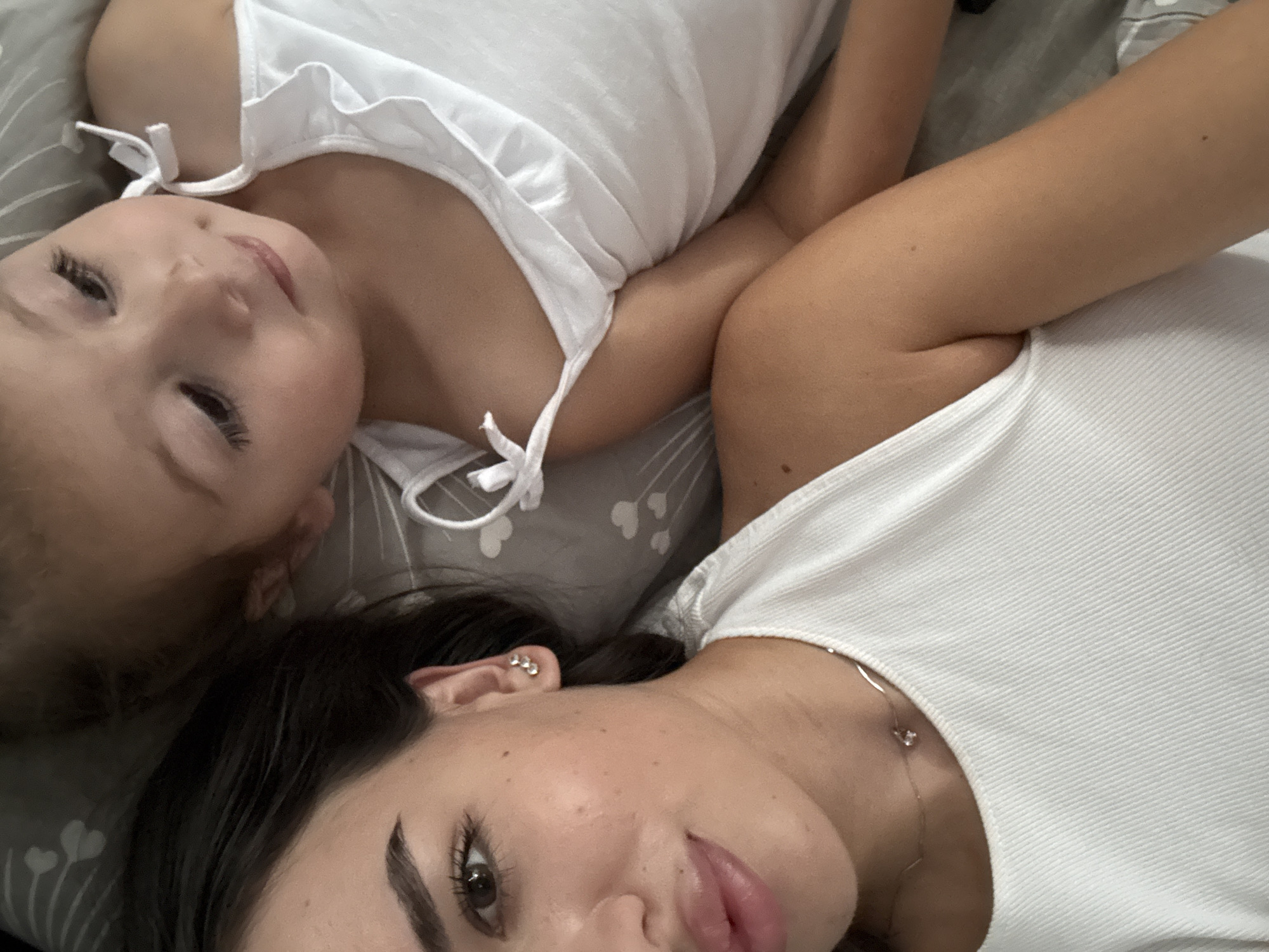 This is a close-up, eye-level, indoor photograph of two females, likely an adult and a young child, lying down. The adult is in the foreground on the left, with dark hair and fair skin. She is wearing a white ribbed tank top and a delicate necklace with a small pendant. Her left ear is visible and adorned with a small, sparkly earring. The child is in the background on the right, with lighter hair and fair skin, wearing a white top with ruffled edges and small bow details on the straps. They are lying on a surface with a gray fabric featuring a white heart-shaped pattern. The lighting suggests an indoor setting, possibly a bedroom. The photograph is taken in Chișinău, Moldova.
